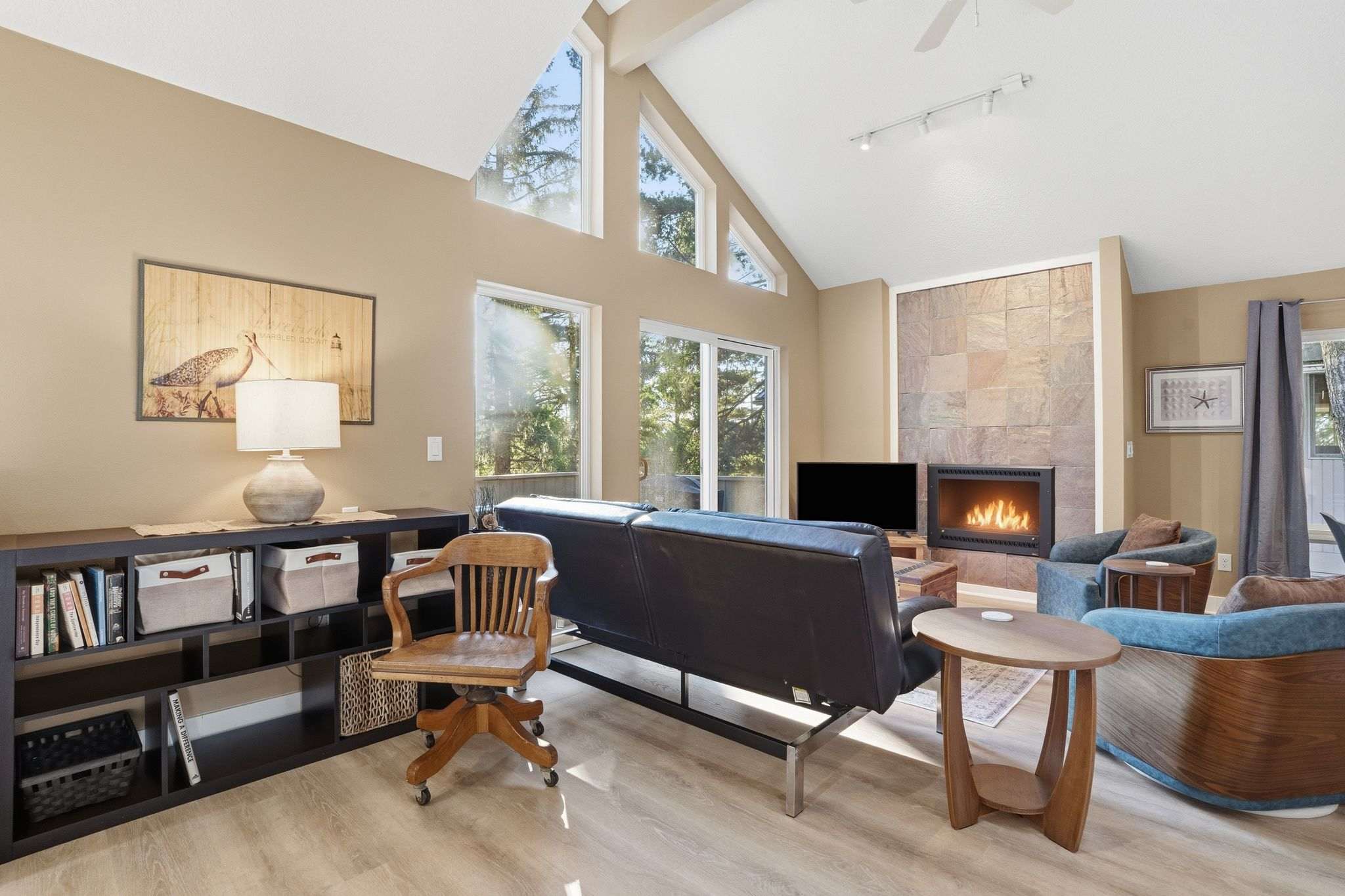 Wide view of the living room with a dark leather sofa, a black bookshelf, and a wooden desk chair, showcasing the vaulted ceiling and large windows that offer views of the surrounding trees.