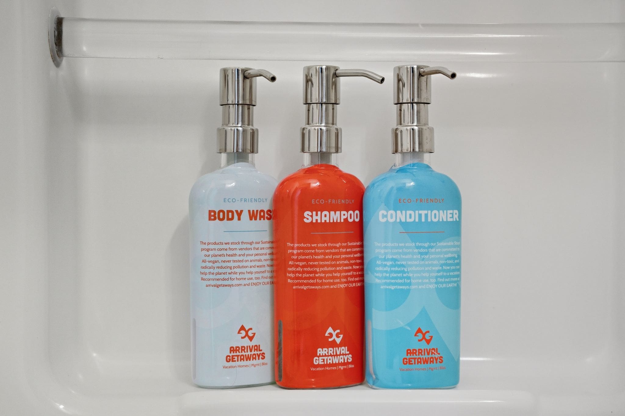 A close-up of three eco-friendly pump bottles, clearly labeled "BODY WASH," "SHAMPOO," and "CONDITIONER" from "ARRIVAL GETAWAYS," provided as bathroom amenities.