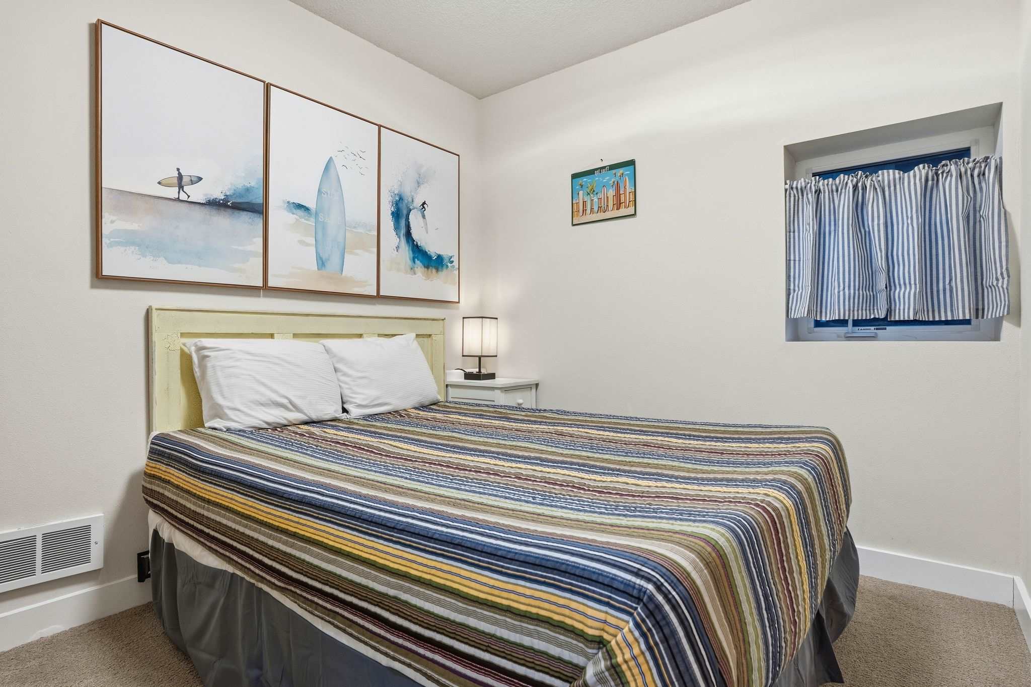 Close-up view of the full-sized bed in bedroom-1, showcasing the striped comforter, light yellow headboard, and surf-themed triptych artwork. A small window with a striped curtain is visible on the right.