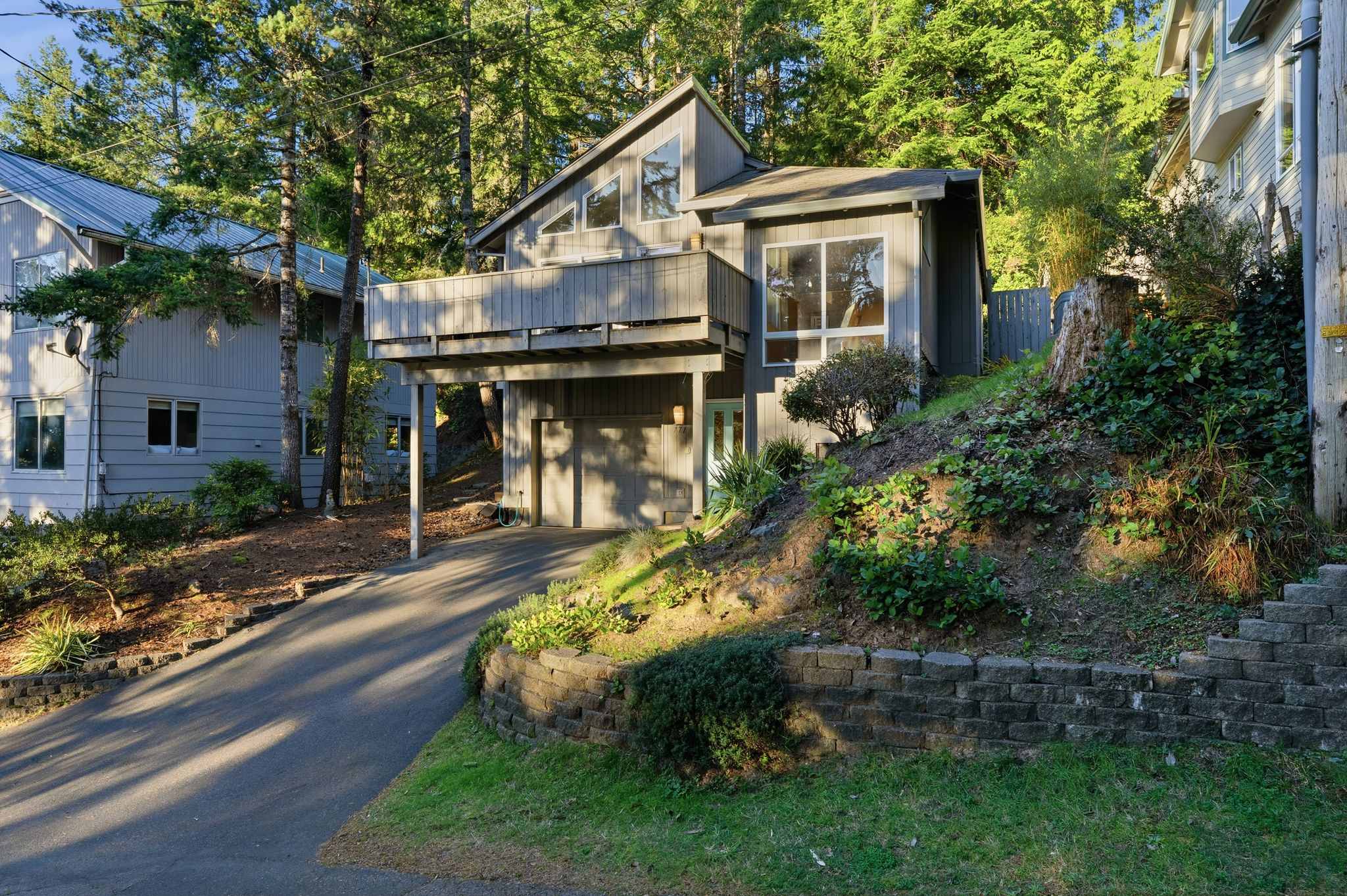 A gray modern house with an elevated balcony and large windows, nestled on a wooded hillside with a paved driveway and a retaining wall, surrounded by tall evergreen trees.
