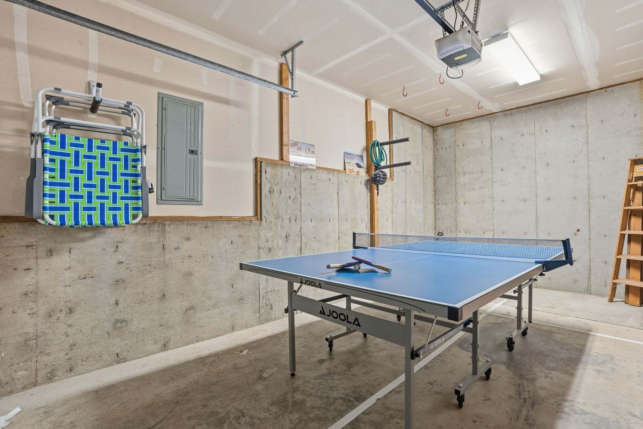 A spacious garage with concrete walls and floor houses a blue Joola ping pong table with paddles and balls. Beach chairs are hung on the wall, and an exposed electrical panel is visible.