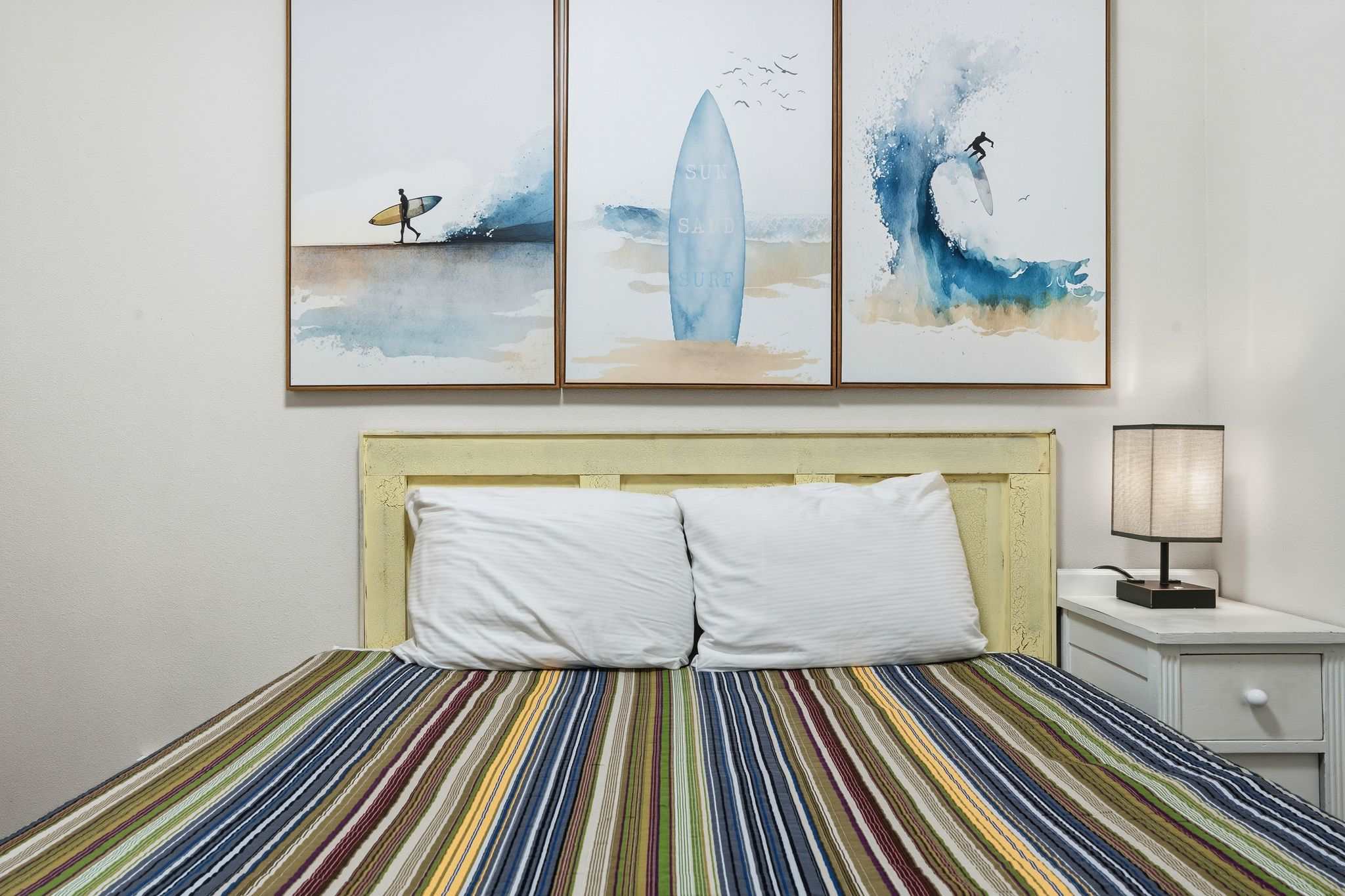 Detailed shot of the full-sized bed in bedroom-1, highlighting the light yellow headboard, white pillows, and the three surf-themed art pieces above. A white nightstand with a lamp is on the right.