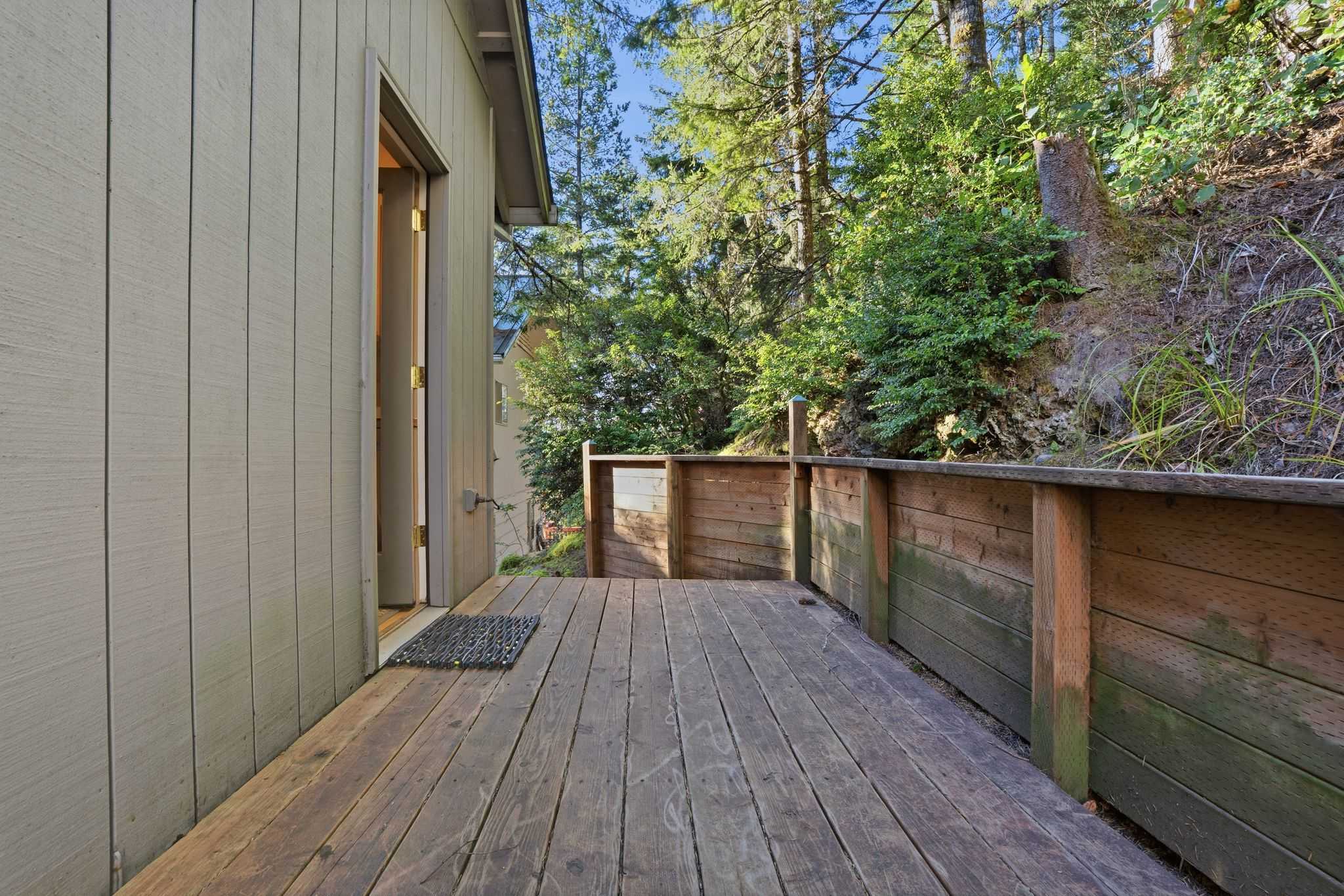 A simple wooden deck area with a low wooden retaining wall, leading to an open doorway, surrounded by dense green foliage and tall trees on a sunny day.