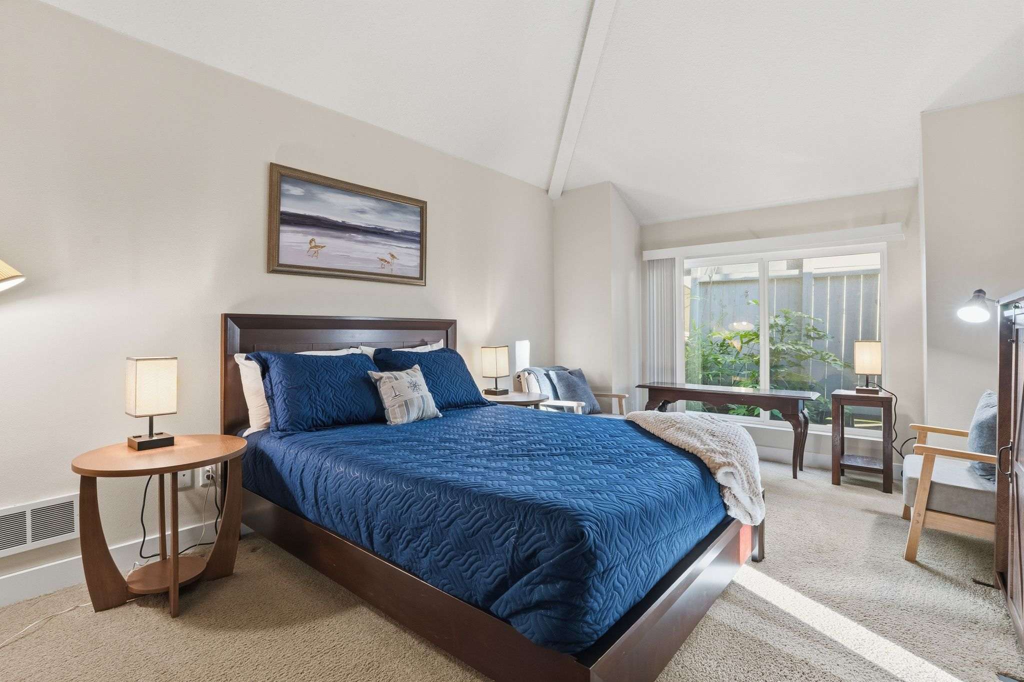 Bright bedroom with a queen-sized bed dressed in blue quilted bedding, flanked by two round wooden nightstands with lamps. A large window provides natural light, and a framed beach painting hangs above the bed.
