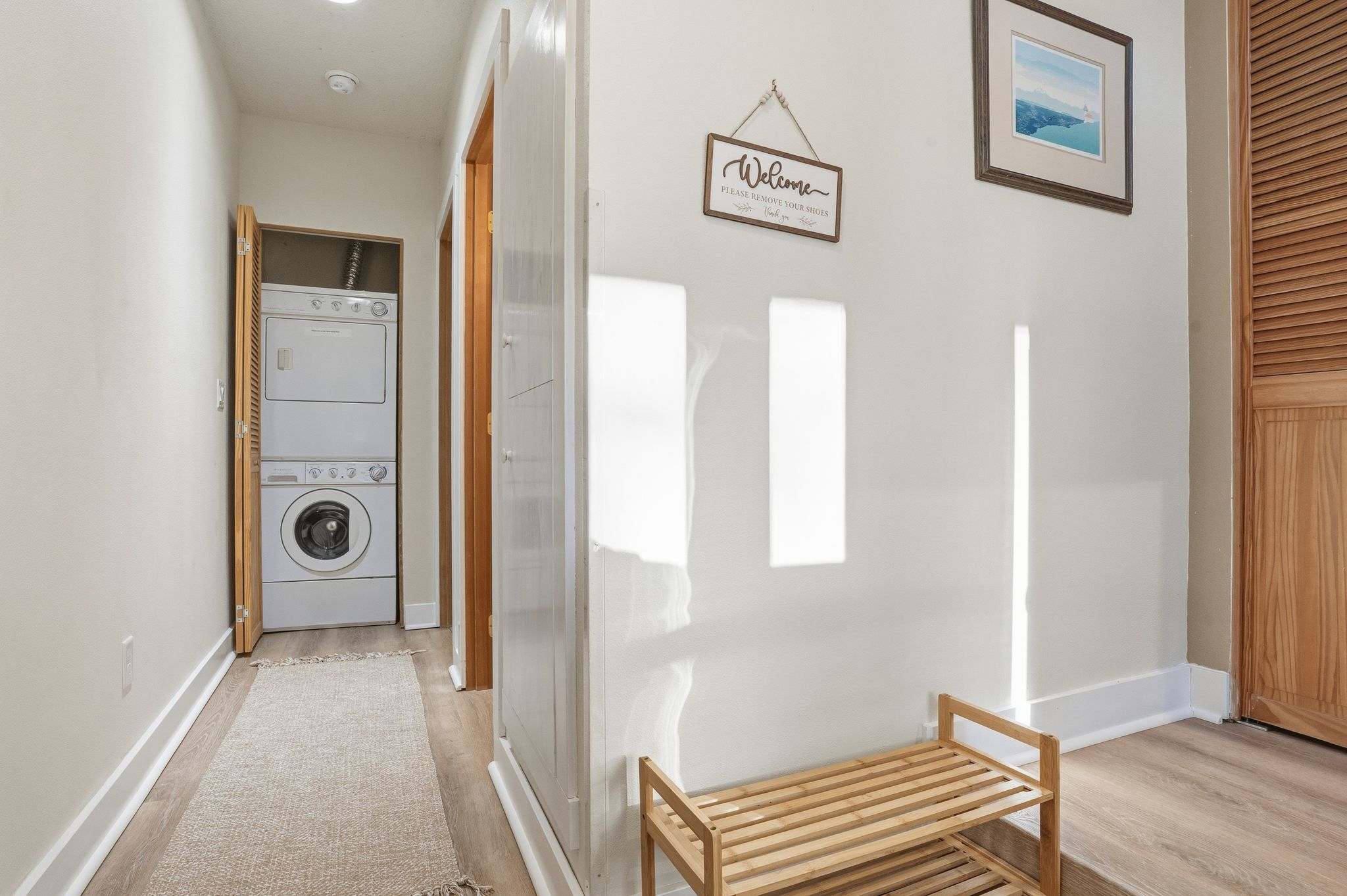 A bright hallway with light wood flooring and a woven rug features a wooden shoe rack and a "Welcome" sign on the wall. A stacked washer and dryer unit is visible in an open closet at the end of the hall.