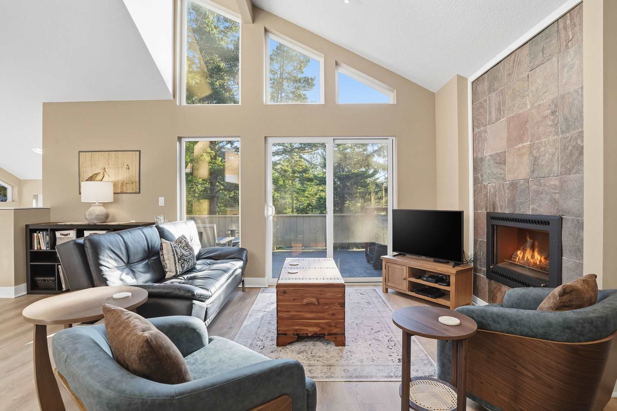 Bright living room with vaulted ceilings, large windows overlooking trees, a dark leather sofa, two blue armchairs, and a stone fireplace with a TV mounted above a wooden media console.