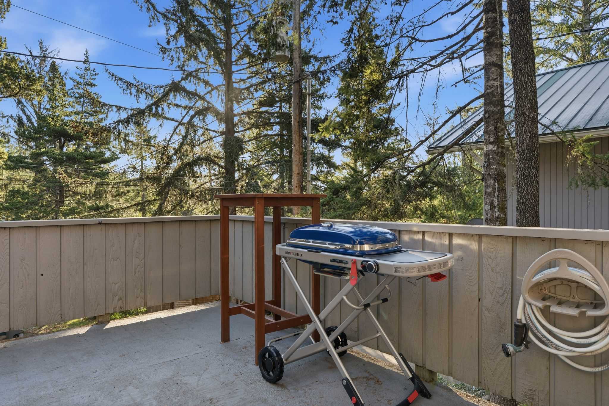 A concrete balcony with a light wood railing, featuring a blue portable BBQ grill on a stand and a tall wooden table, offering a view of tall evergreen trees.