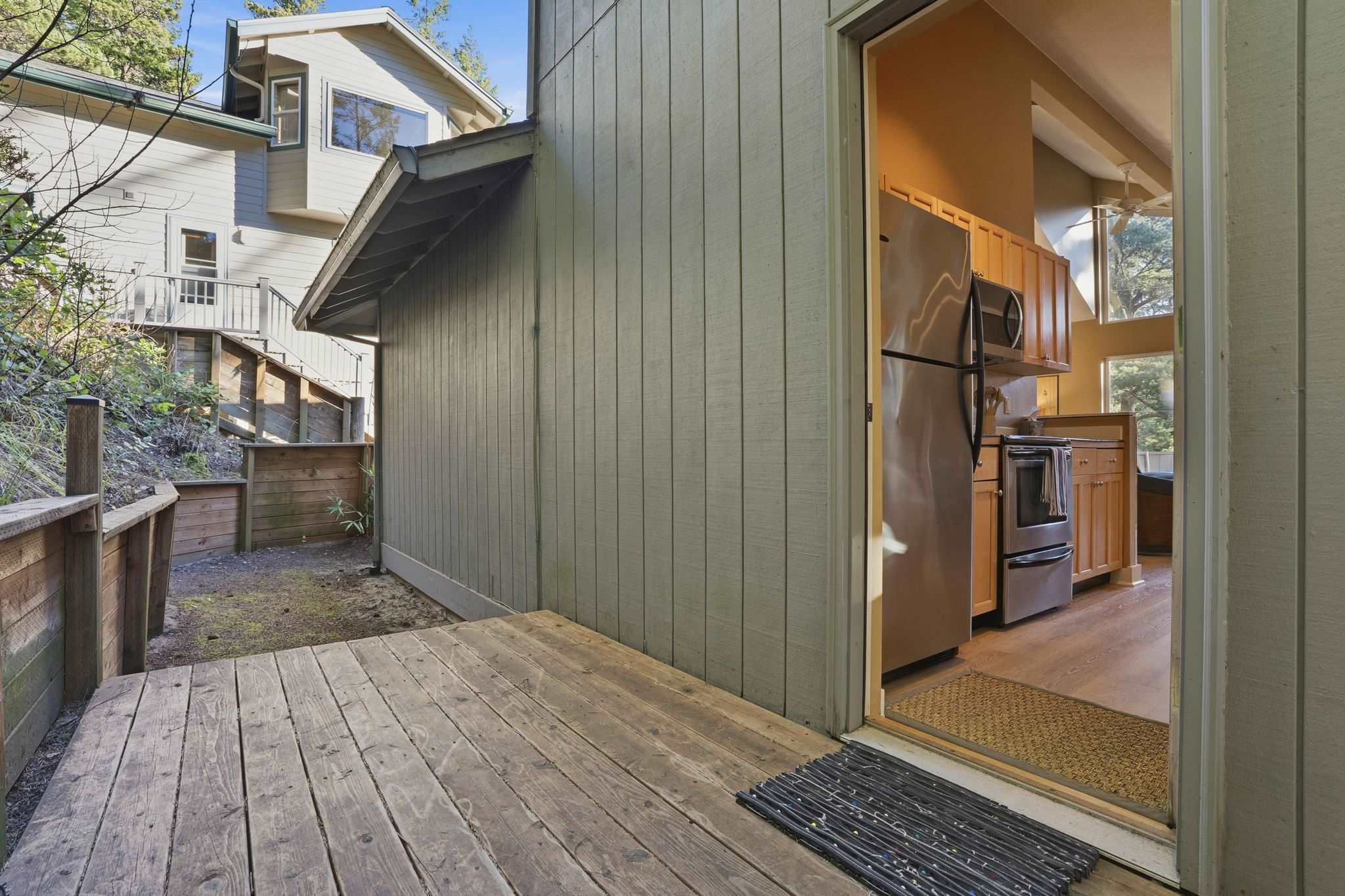 View of a wooden deck leading to the kitchen entrance, with a light green-sided house exterior and lush green foliage in the background.