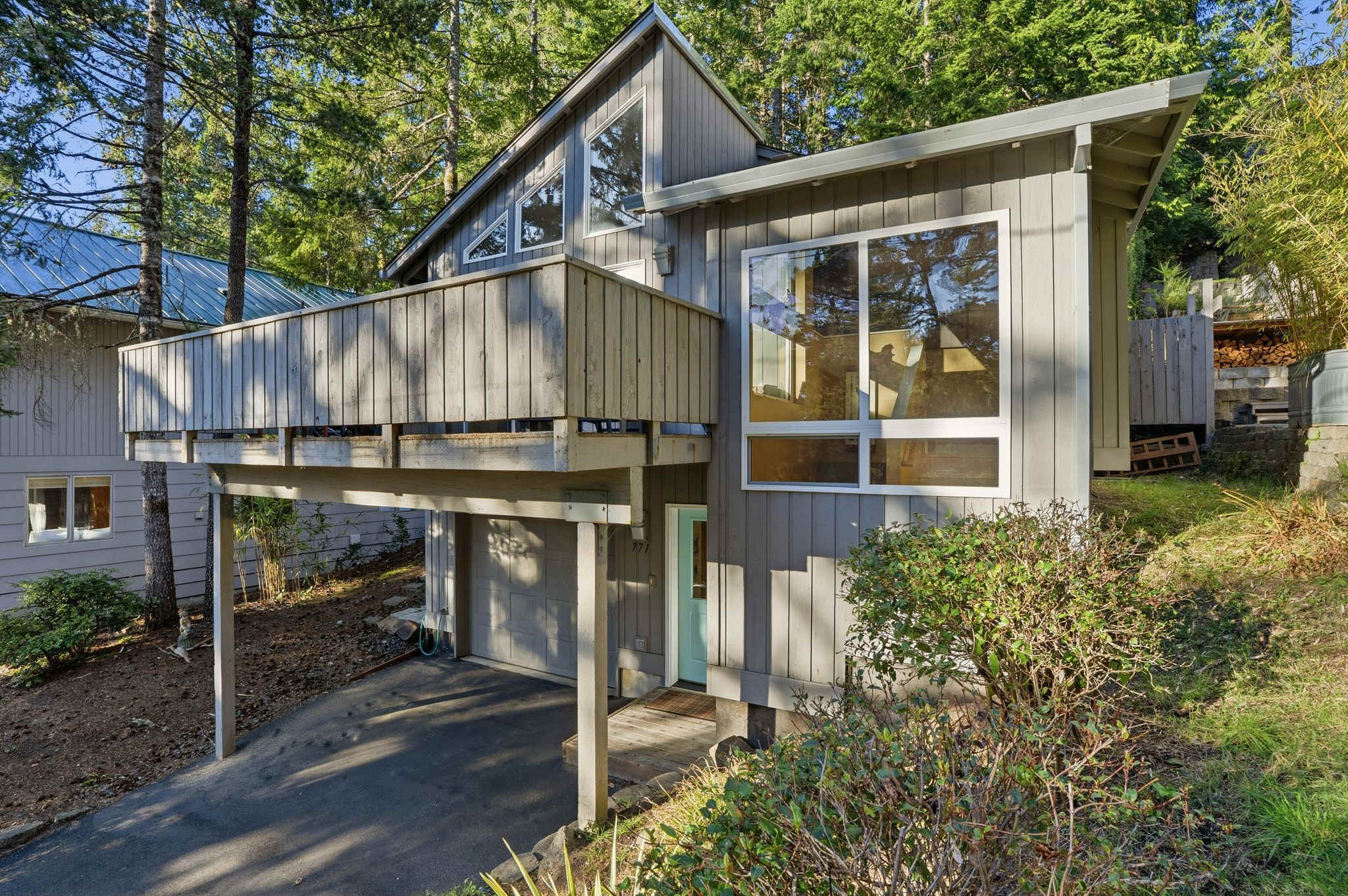 A gray modern house with an elevated balcony and large windows, surrounded by dense evergreen trees, featuring a sloped driveway leading to a garage and a light blue front door.