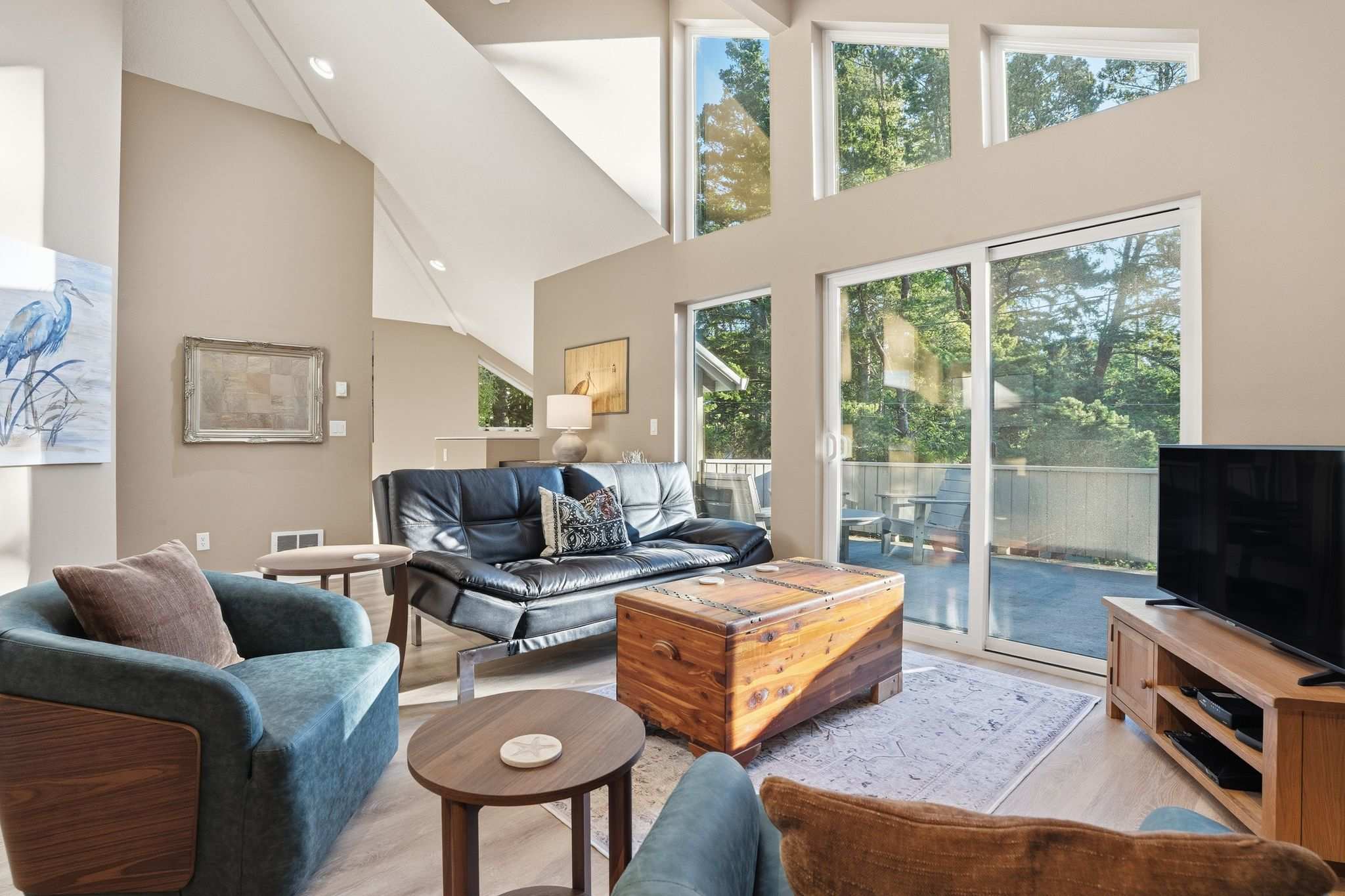 Spacious living room featuring a dark leather sofa, two blue armchairs, a wooden chest coffee table, and large windows with a sliding glass door leading to an outdoor deck, all under a vaulted ceiling.