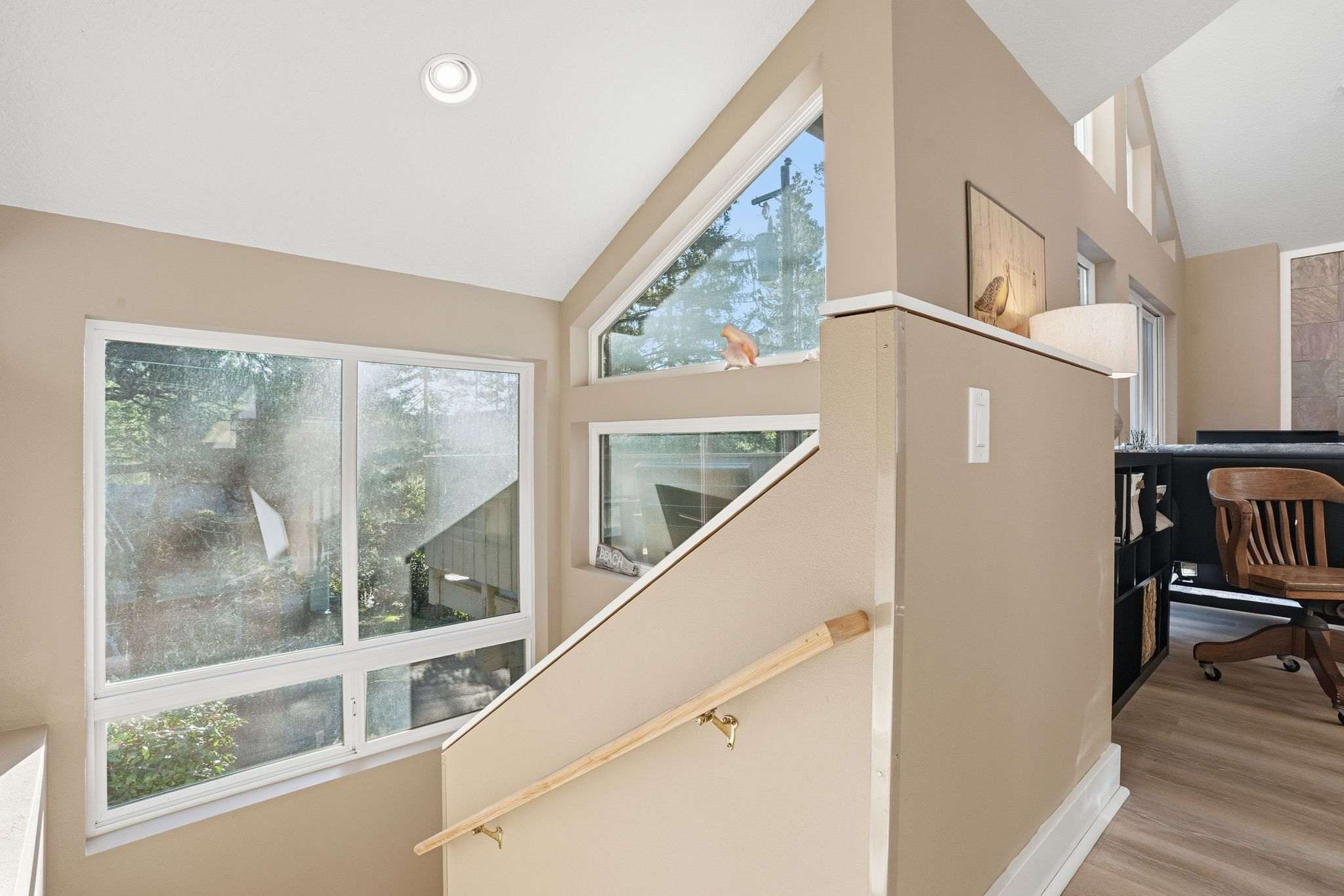 A modern staircase with light wood handrail and beige walls features large, irregularly shaped windows offering views of lush greenery outside. The area is brightly lit with natural light and recessed ceiling lights.