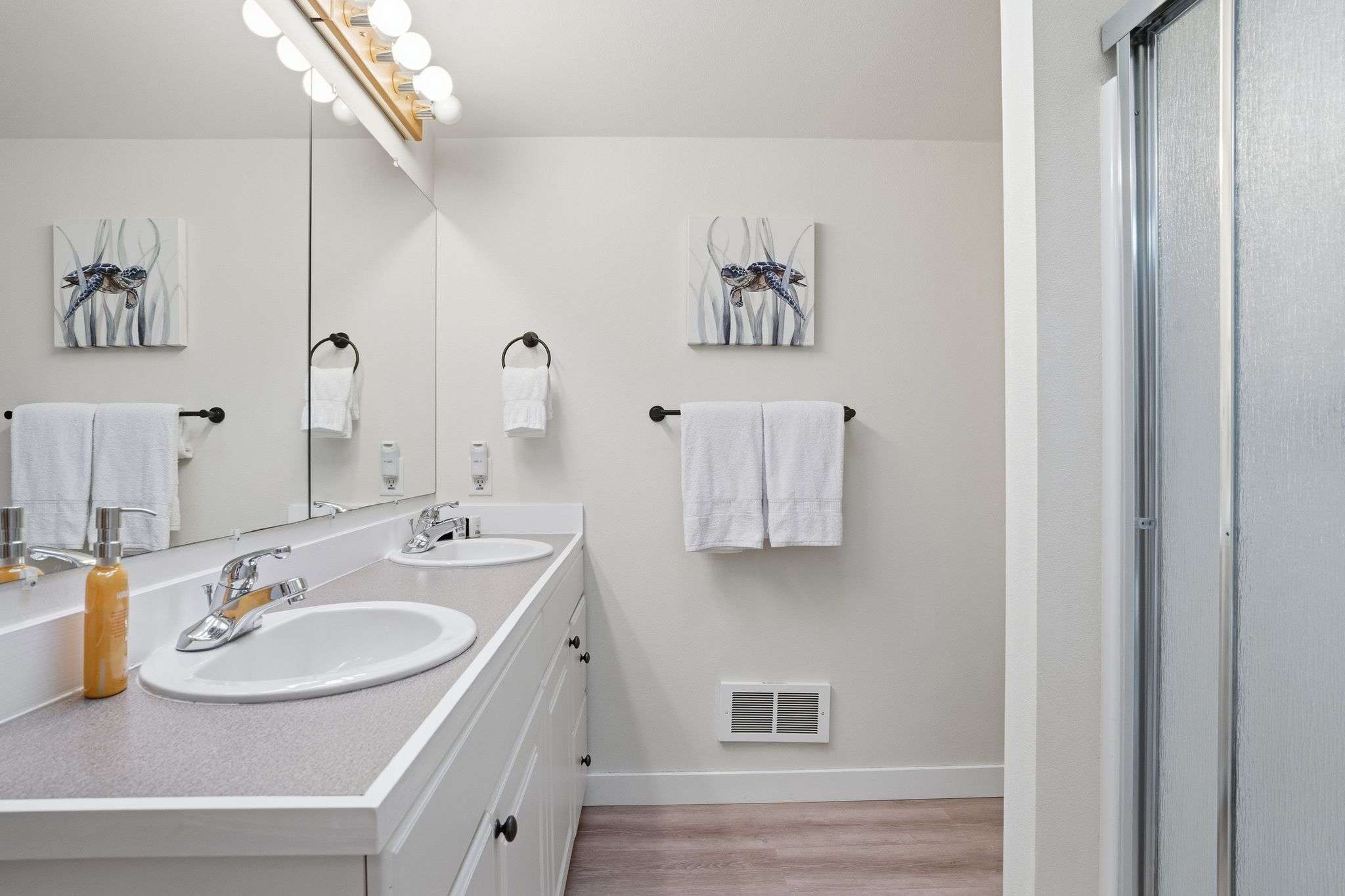 A bright bathroom features a double vanity with two sinks, a large mirror, and a separate shower stall with a frosted glass door. Two framed artworks depicting blue sea turtles adorn the light-colored wall.
