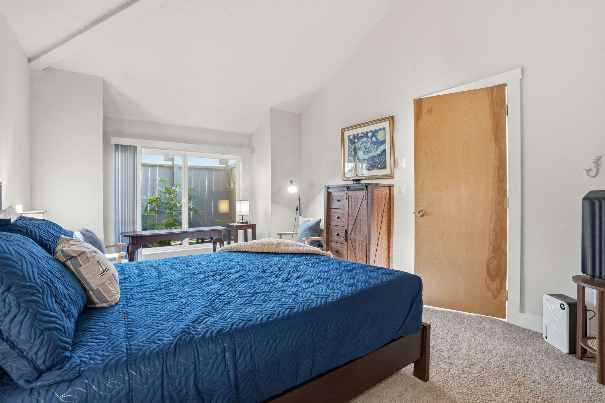 Wide view of bedroom-2, featuring a queen-sized bed with blue bedding, a dark wood dresser, and a large window looking out onto a private outdoor area. A small seating area with a chair and table is visible near the window.