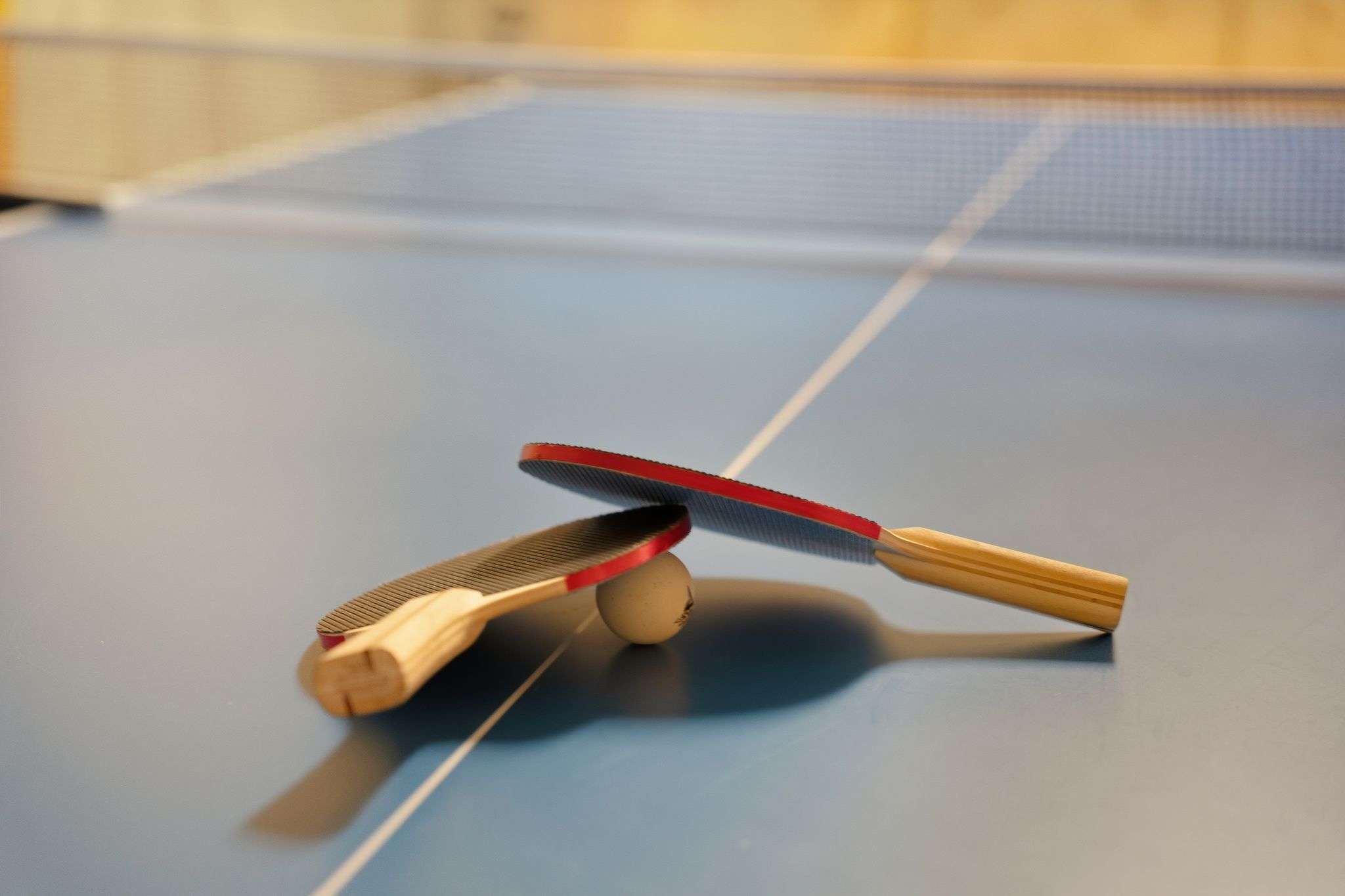 A close-up shot of two red and black ping pong paddles and a white ball resting on a blue ping pong table, with the white net visible in the background.