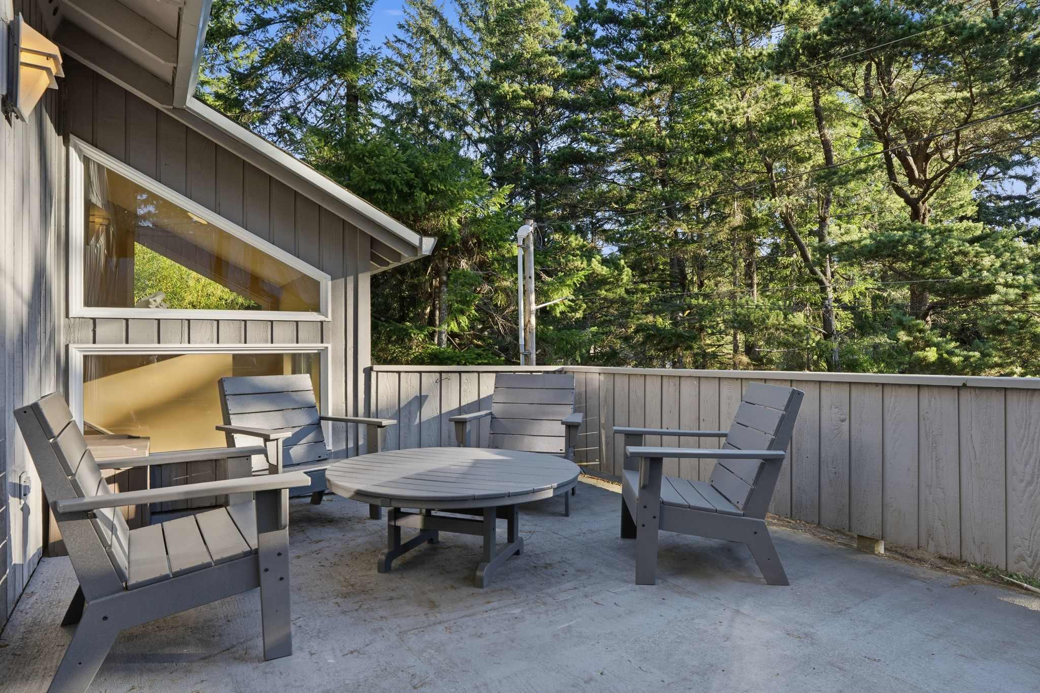 An outdoor concrete deck area with light gray Adirondack-style chairs and a matching round table, surrounded by a light wood fence and dense evergreen trees.