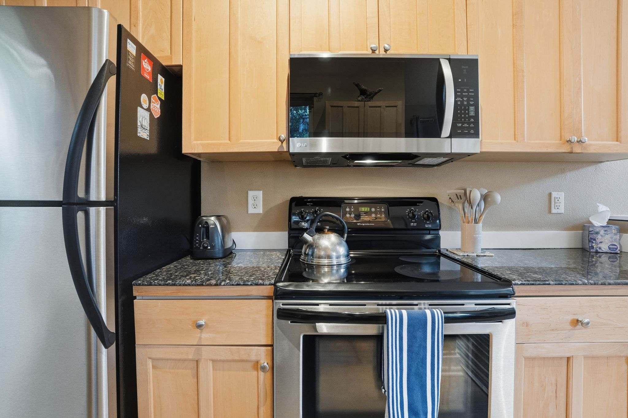 Close-up of the kitchen's cooking area, featuring a stainless steel refrigerator, black electric stove, and microwave, all surrounded by light wood cabinetry and dark granite countertops.