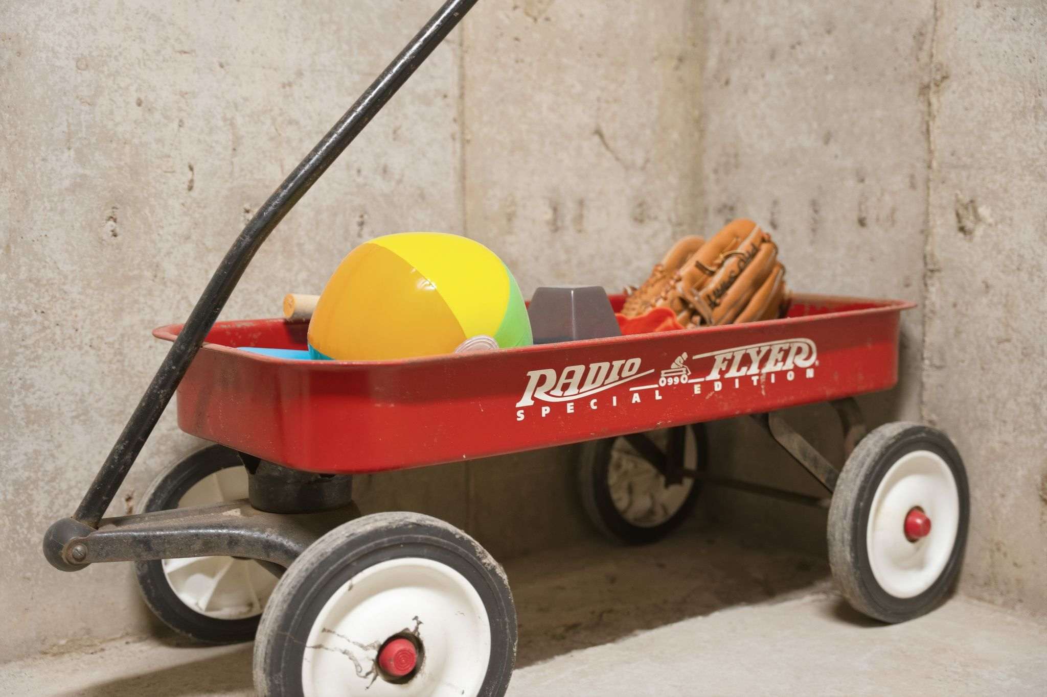 A classic red Radio Flyer wagon, labeled "Special Edition," is filled with various recreational items including a colorful beach ball, a baseball glove, and a small bucket, set against a concrete wall.