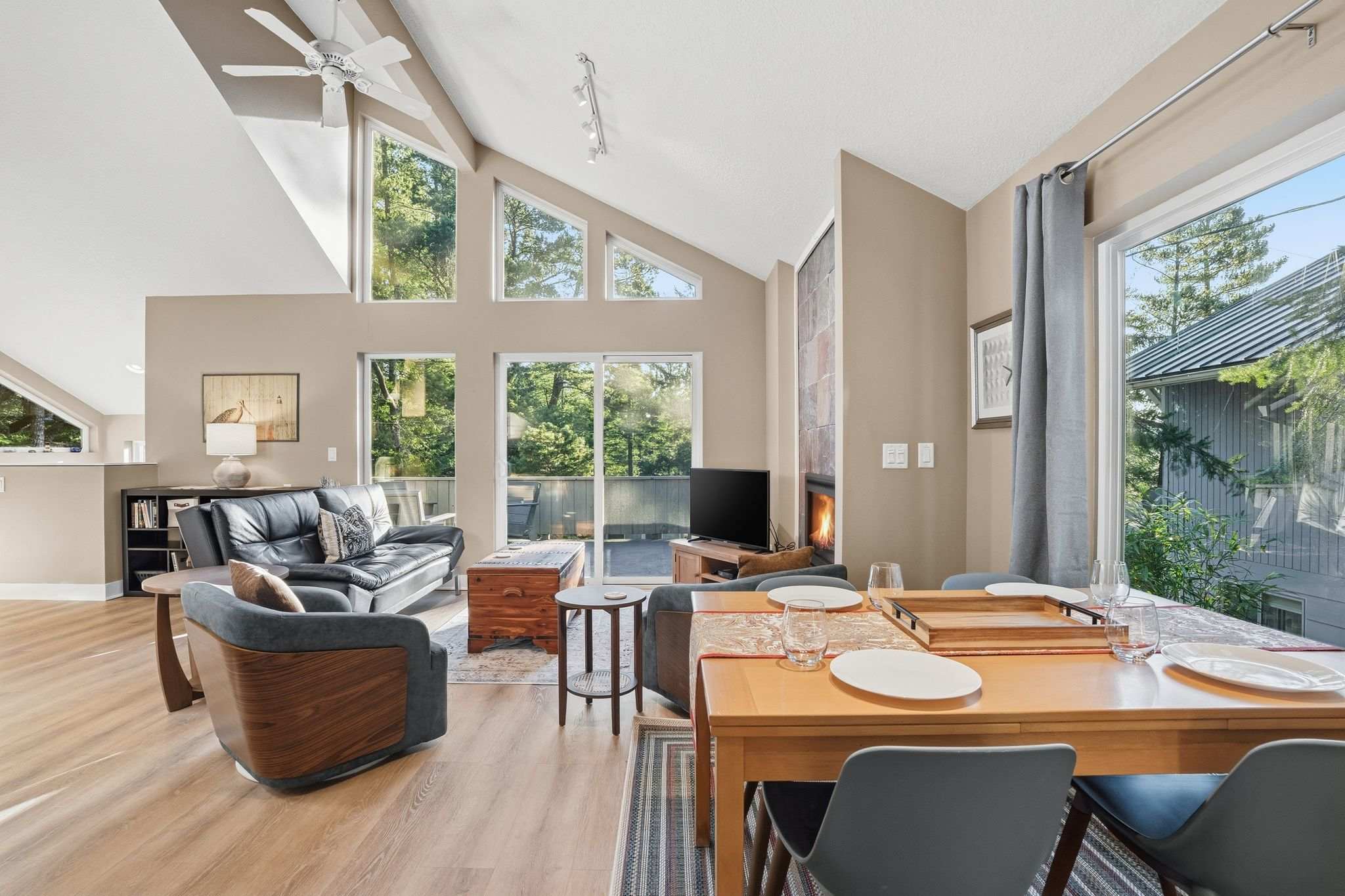 Bright, open-concept living and dining area with vaulted ceilings, large windows overlooking trees, a modern fireplace, and light wood flooring. The living space features a black leather sofa and a dark wood armchair, while the dining area has a light wood table with blue chairs.
