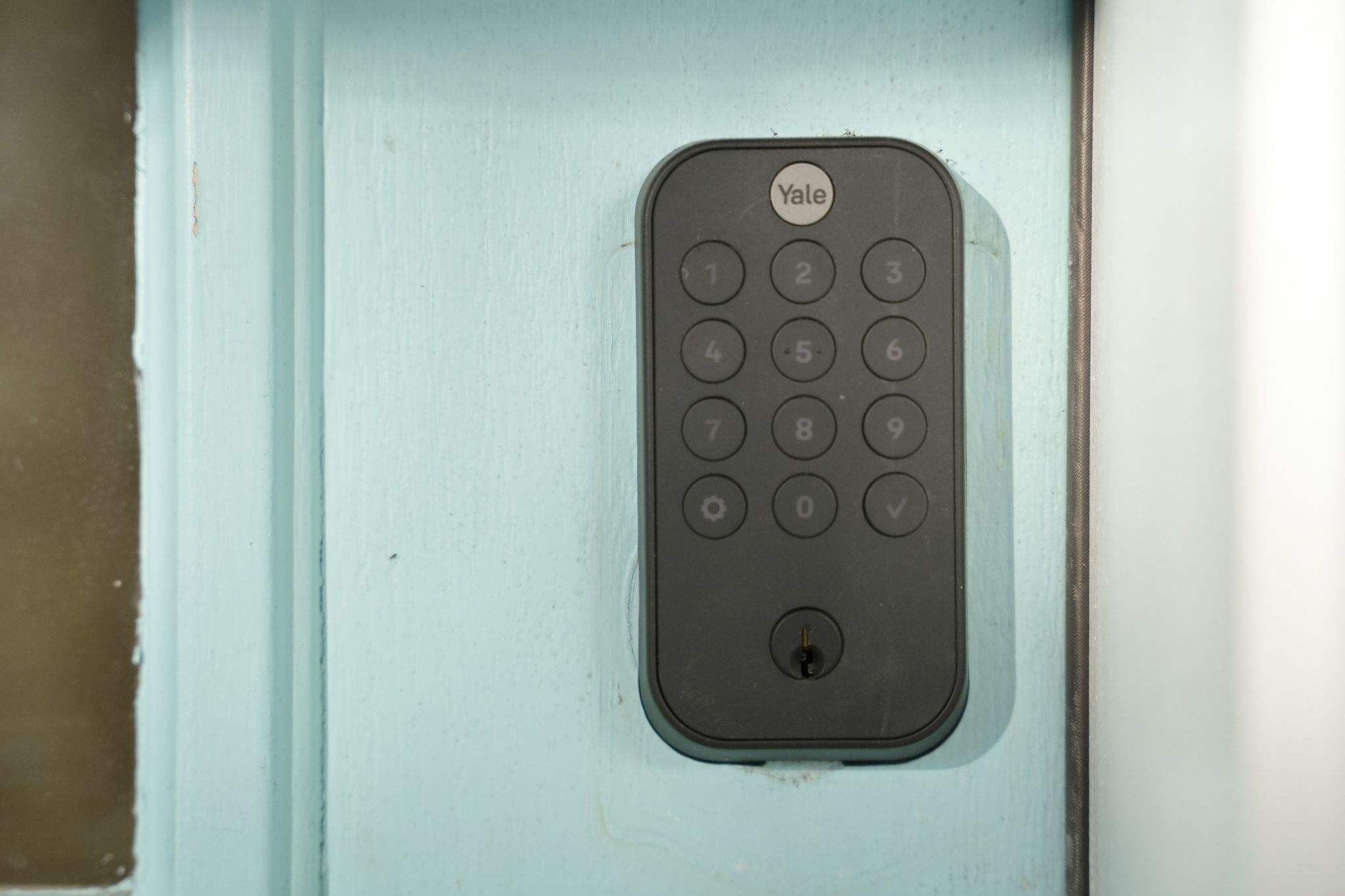 A close-up of a dark gray Yale smart lock with a keypad and keyhole, mounted on a light blue wooden door. The door has visible wood grain and a slightly distressed paint finish.