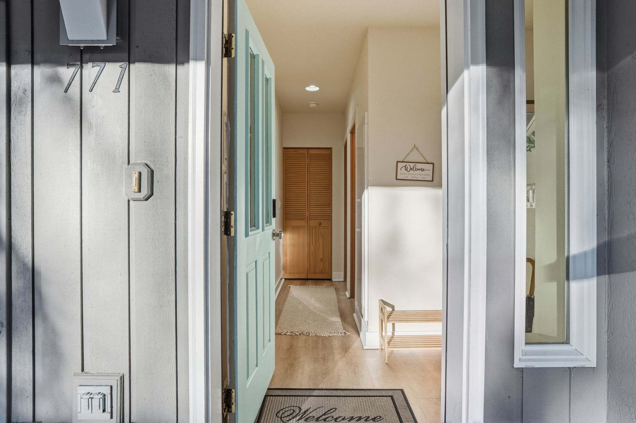 The light blue front door is open, revealing a welcoming entryway with light wood flooring and a "Welcome" mat. A wooden shoe rack and a "Welcome" sign hang on the wall, leading to a hallway with a closet.