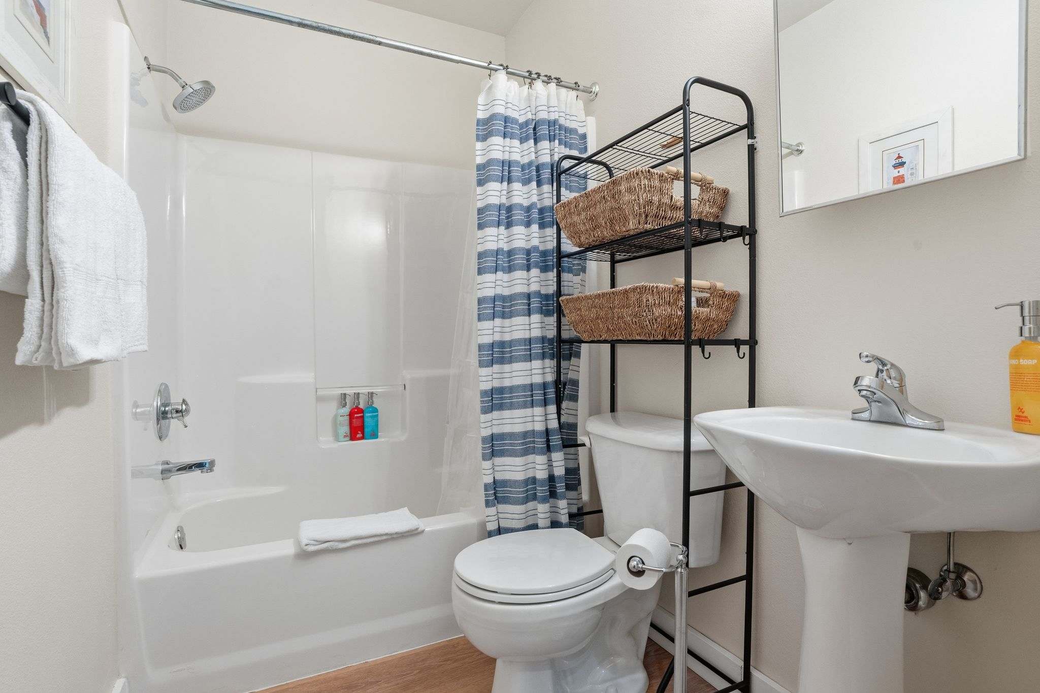 A bright bathroom features a white tub/shower combo with a blue and white striped curtain, a white toilet, and a pedestal sink. A black metal shelving unit above the toilet holds woven baskets.