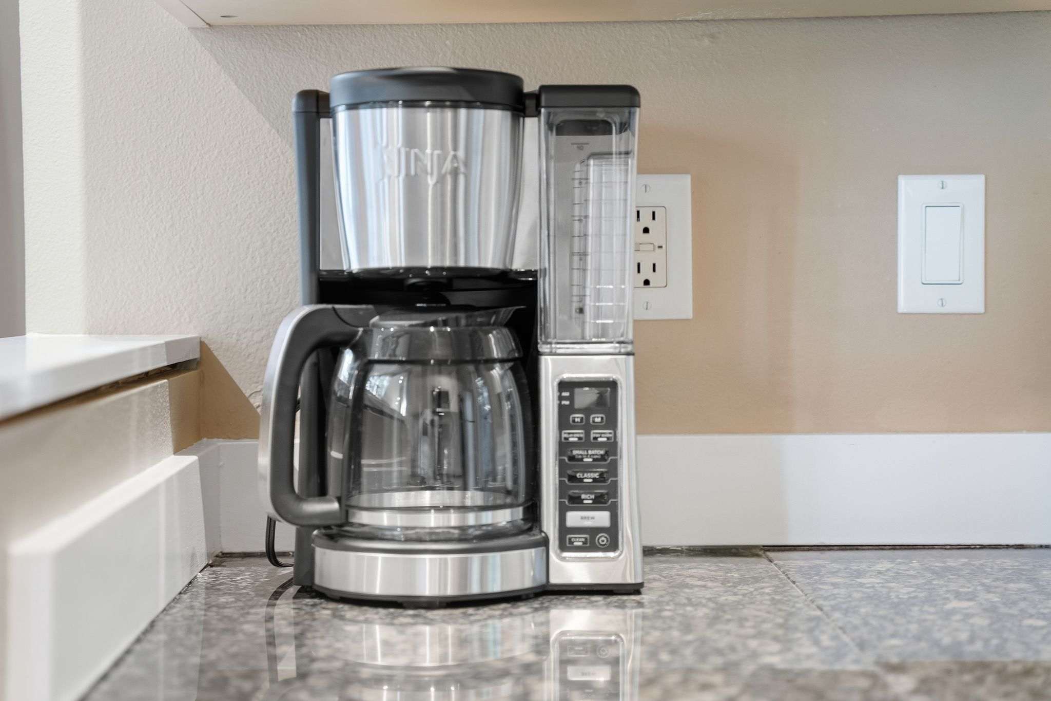 A stainless steel and black Ninja coffee maker sits on a speckled granite countertop in a kitchen. A white electrical outlet and light switch are visible on the beige wall behind it.
