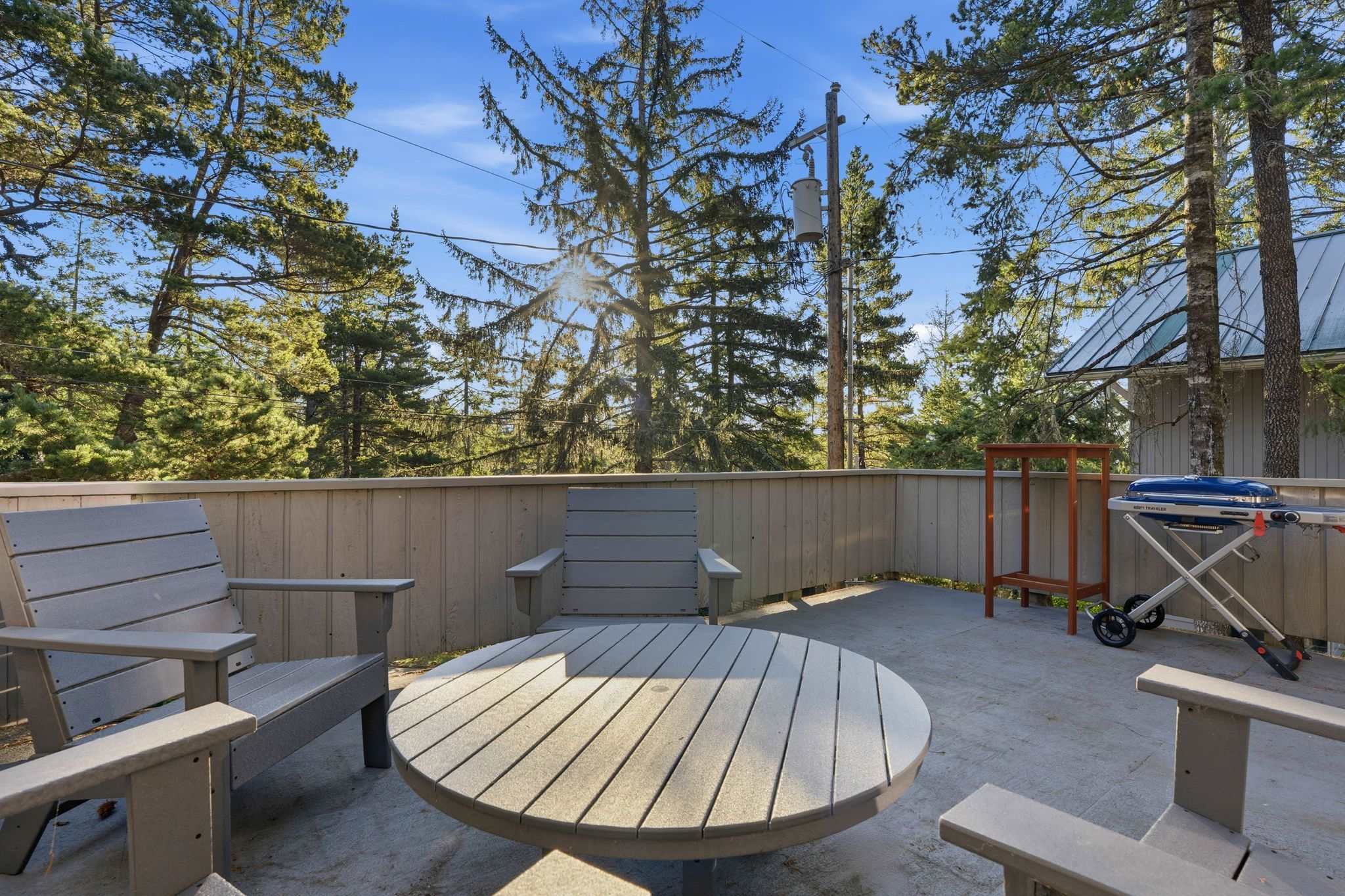 Outdoor deck with light gray slatted furniture, including a round table and chairs, surrounded by a wooden fence and tall pine trees under a bright blue sky. A BBQ grill is visible in the background.