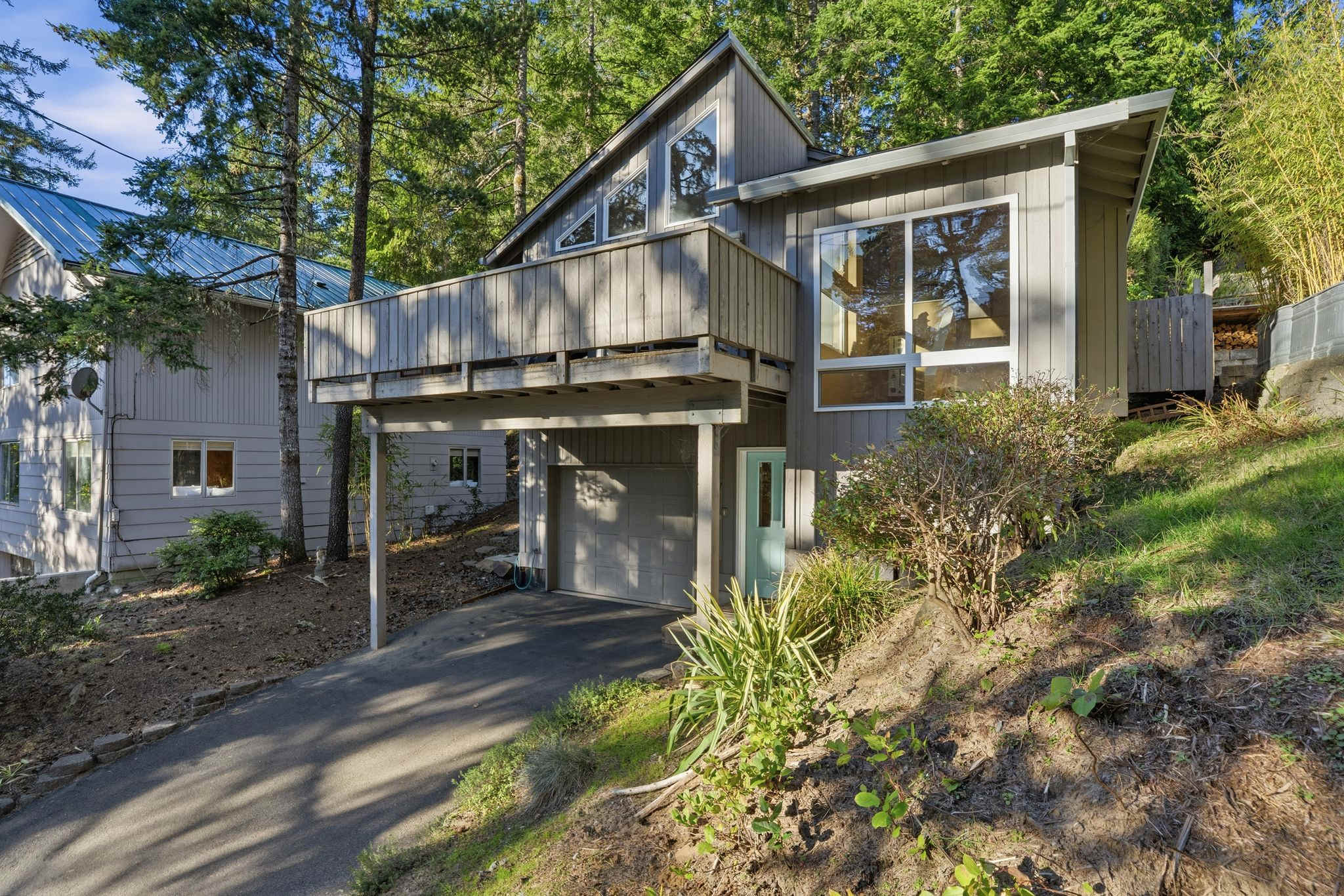 A gray modern house with an elevated balcony and large windows, situated on a sloped, wooded lot with a paved driveway leading to a garage, surrounded by tall evergreen trees.