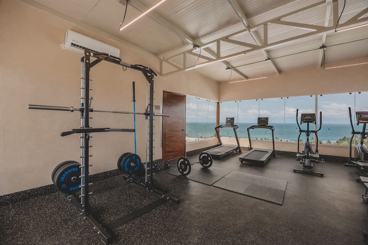 Well-equipped gym with rubber flooring, featuring a power rack with weights, treadmills, and an elliptical machine, all positioned to offer expansive ocean views through large windows. The ceiling has exposed white trusses.