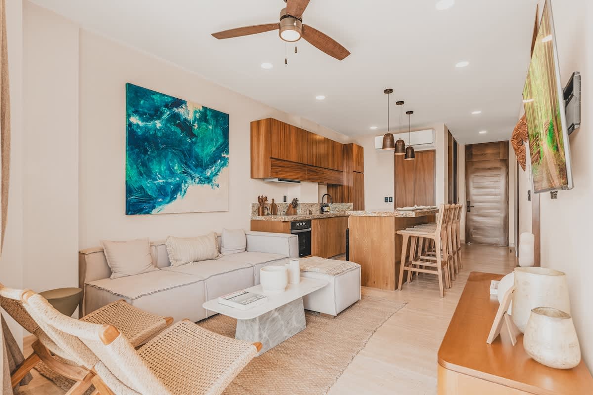 Open-concept living area with a light-colored sectional sofa, a modern white coffee table, and a textured rug, seamlessly connecting to the kitchen with wooden cabinetry and granite island.