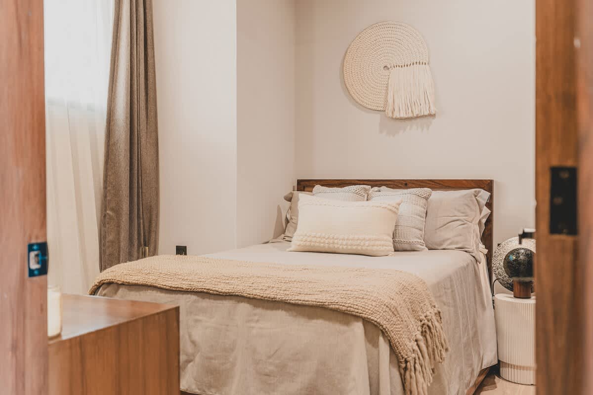 A cozy second bedroom features a queen-sized bed with neutral bedding and a wooden headboard, adorned with a round woven wall hanging above. Light-colored curtains frame a window on the left.