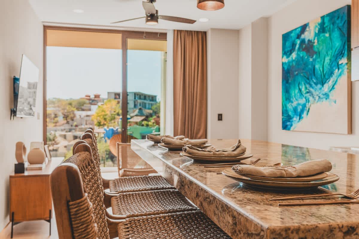 A spacious dining area features a long granite-topped island with several woven bar stools, set against a backdrop of large windows and a vibrant blue abstract painting. A ceiling fan is visible overhead.