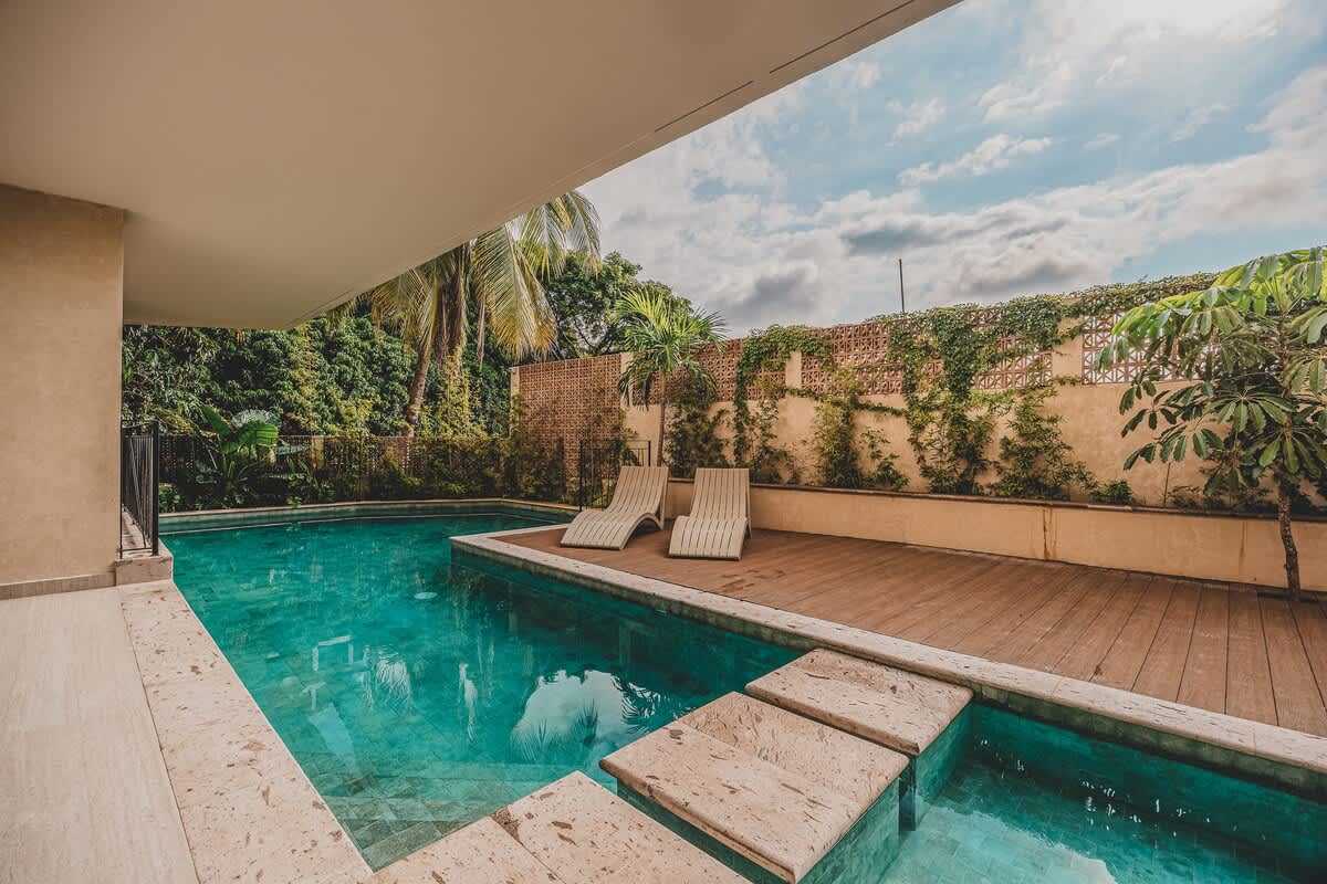 A serene outdoor swimming pool with turquoise water and stepping stones leads to a wooden deck with two lounge chairs, bordered by a textured wall covered in climbing plants and palm trees.