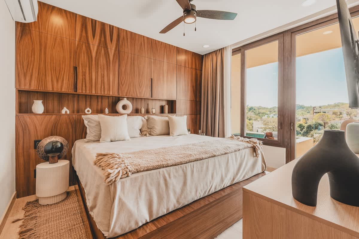A modern bedroom featuring a large bed with light bedding and a throw, set against a striking dark wood paneled wall with integrated shelving, and a balcony with a view.