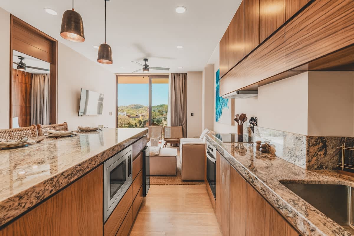 A modern galley kitchen features light wooden cabinetry, speckled granite countertops, and stainless steel appliances, with a microwave built into the island and a view towards the open-concept living area.