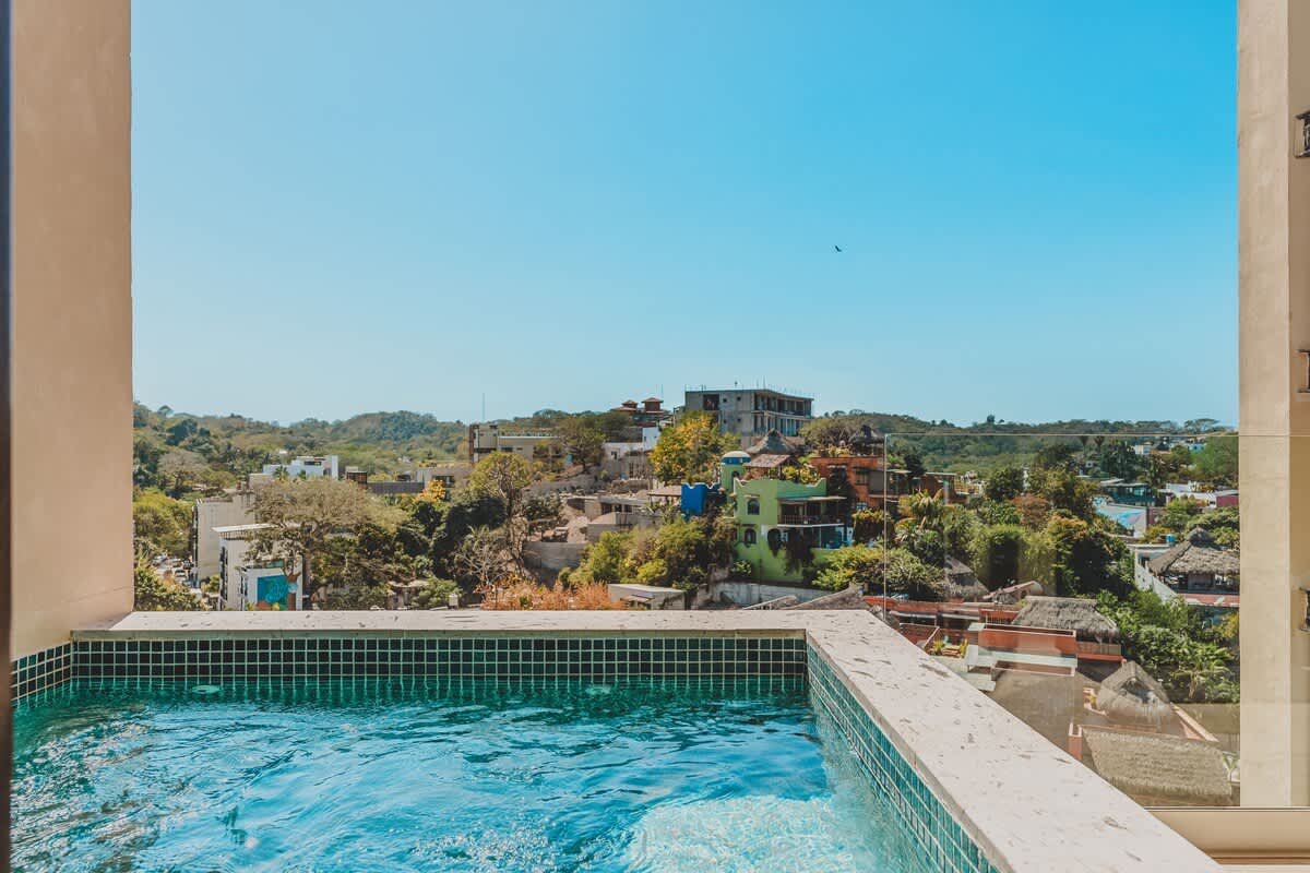 A private plunge pool or hot tub with blue mosaic tiles, offering a panoramic view of a lush, green town and distant hills under a clear blue sky.