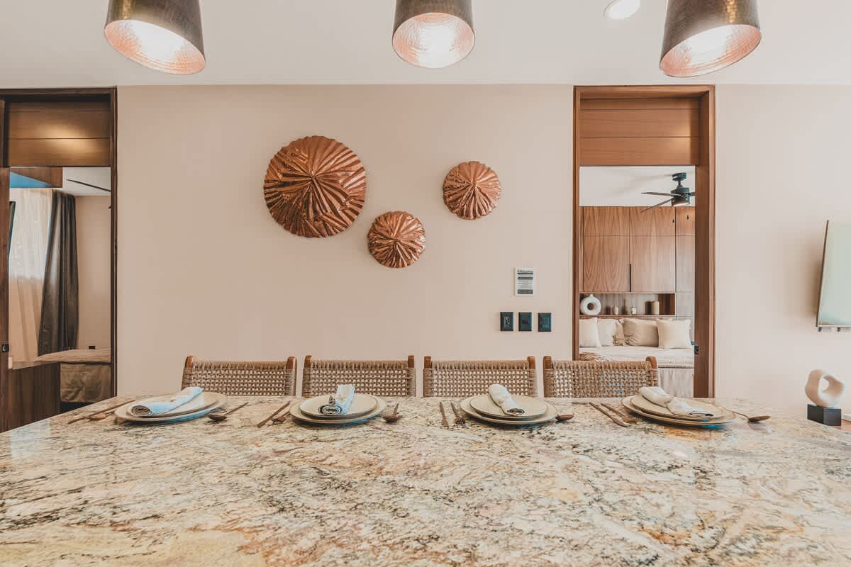 The dining area features a large granite-topped kitchen island set with four place settings, complemented by three copper pendant lights and decorative copper wall art, with a glimpse of the living room beyond.