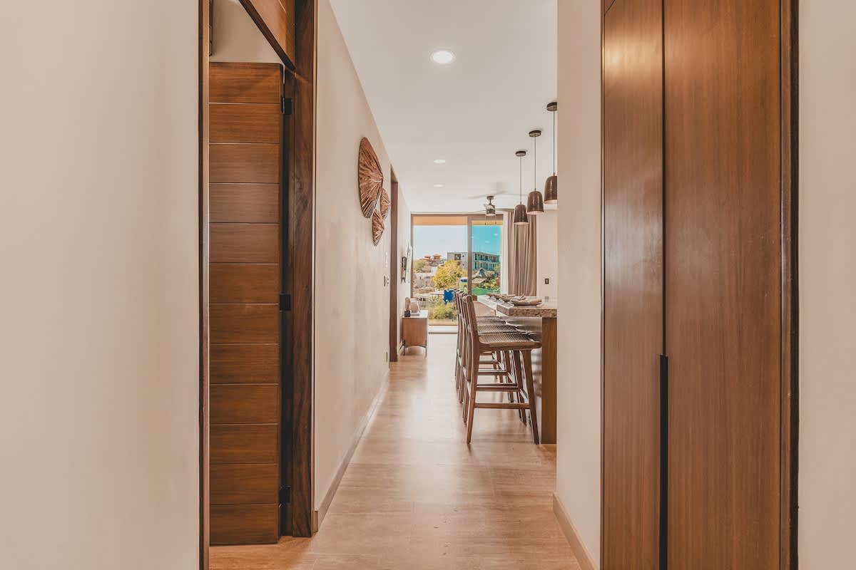 A bright hallway with light tile flooring and neutral walls leads past a wooden built-in cabinet and decorative wall art, opening into the main living space with a view of the kitchen island and balcony.