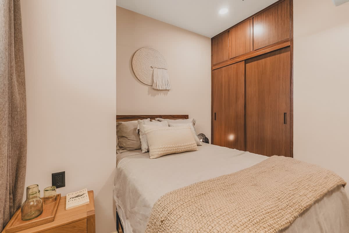 Close-up view of the second bedroom's bed with neutral bedding and a knitted throw, a circular woven wall hanging, and a wooden nightstand with decorative items.