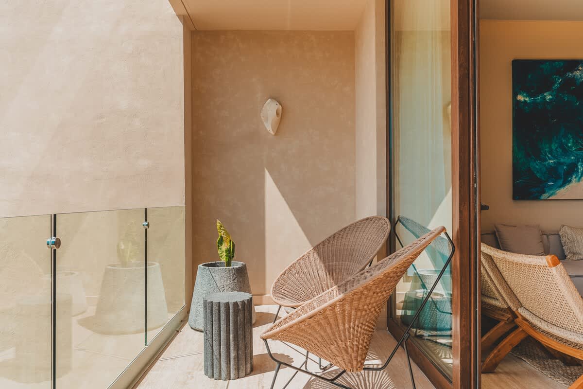 A sunlit balcony features two woven wicker chairs and a cylindrical side table, with a potted plant and a glass railing offering an open view. The wall has a unique shell-like decorative piece.