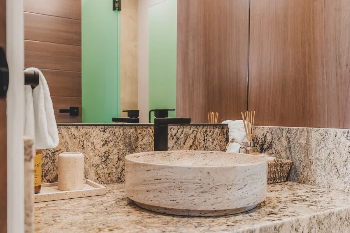 A modern bathroom features a round, light-colored stone vessel sink set on a granite countertop with a black faucet. A large mirror reflects a green-tinted shower enclosure and wooden wall paneling.