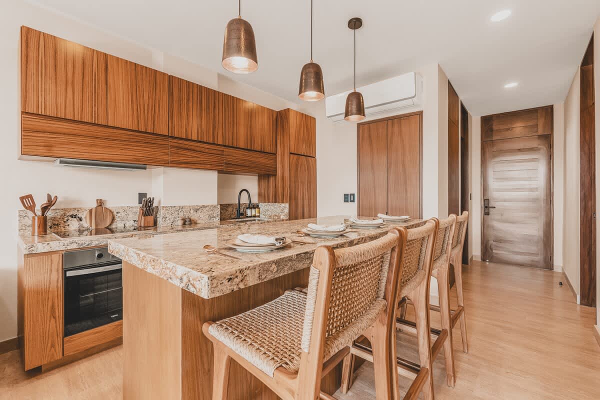 Modern kitchen featuring rich wooden cabinetry, speckled granite countertops, and a large island with four woven bar stools, illuminated by three copper pendant lights.