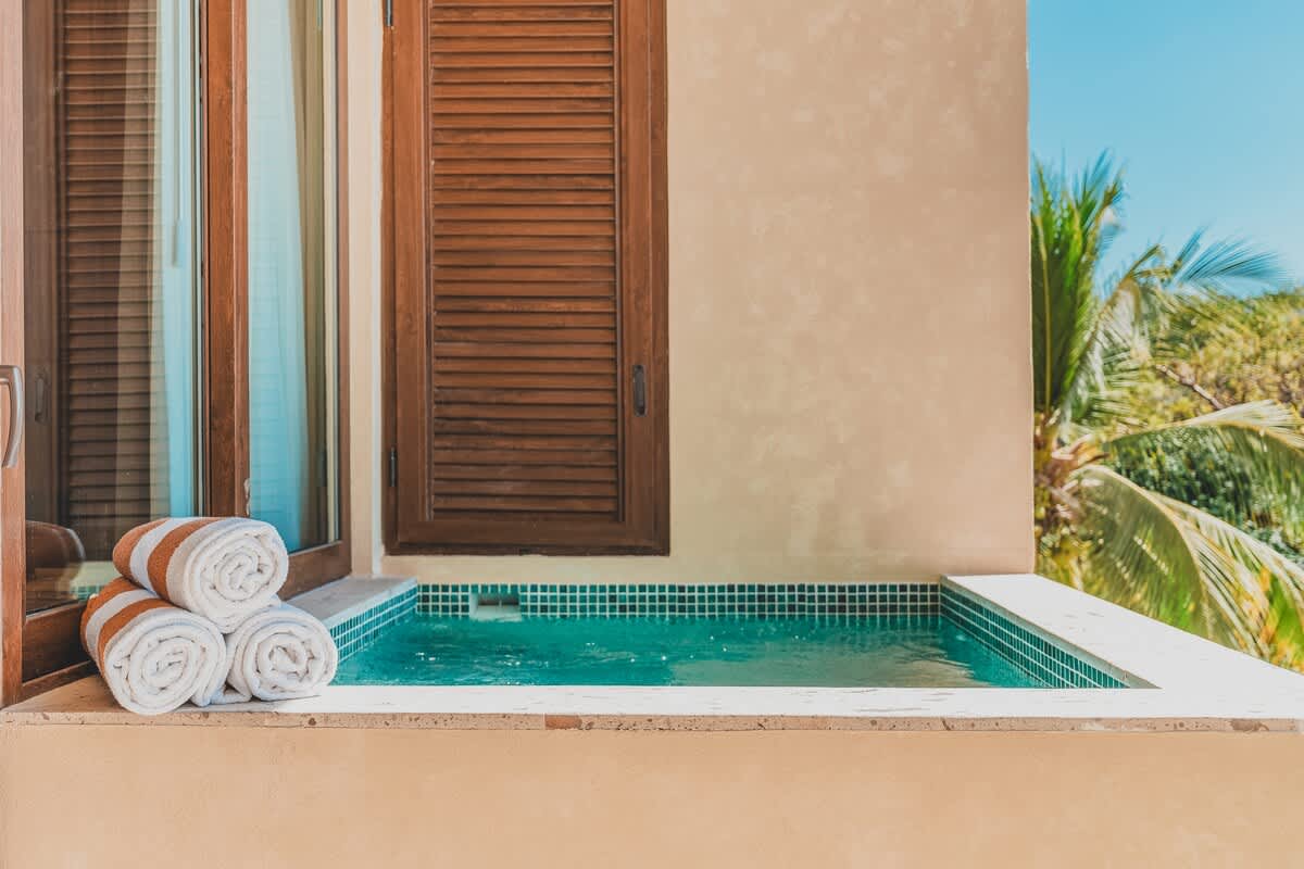 A private outdoor plunge pool or hot tub with turquoise mosaic tiles, located on a balcony next to a window with wooden shutters, and three neatly rolled towels.