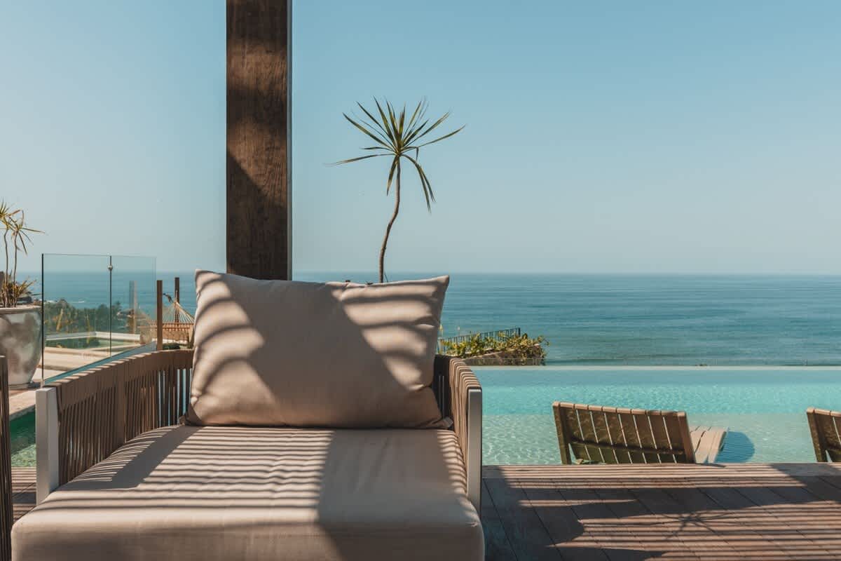 A comfortable outdoor lounge chair with a beige cushion sits on a wooden deck, offering a serene view of the sparkling blue ocean and an infinity pool edge under a clear sky, with a small palm tree in the foreground.