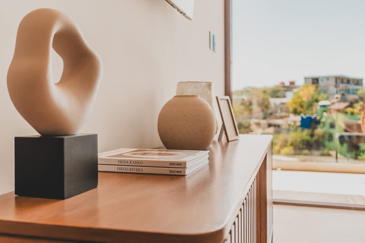 A stylish wooden console table adorned with a modern abstract sculpture, ceramic vases, framed art, and books, with a bright window offering a view of the outdoors.