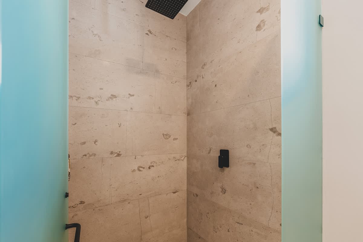 A modern shower with light beige large-format tile walls, featuring a black rain shower head and black minimalist controls, enclosed by frosted glass panels.
