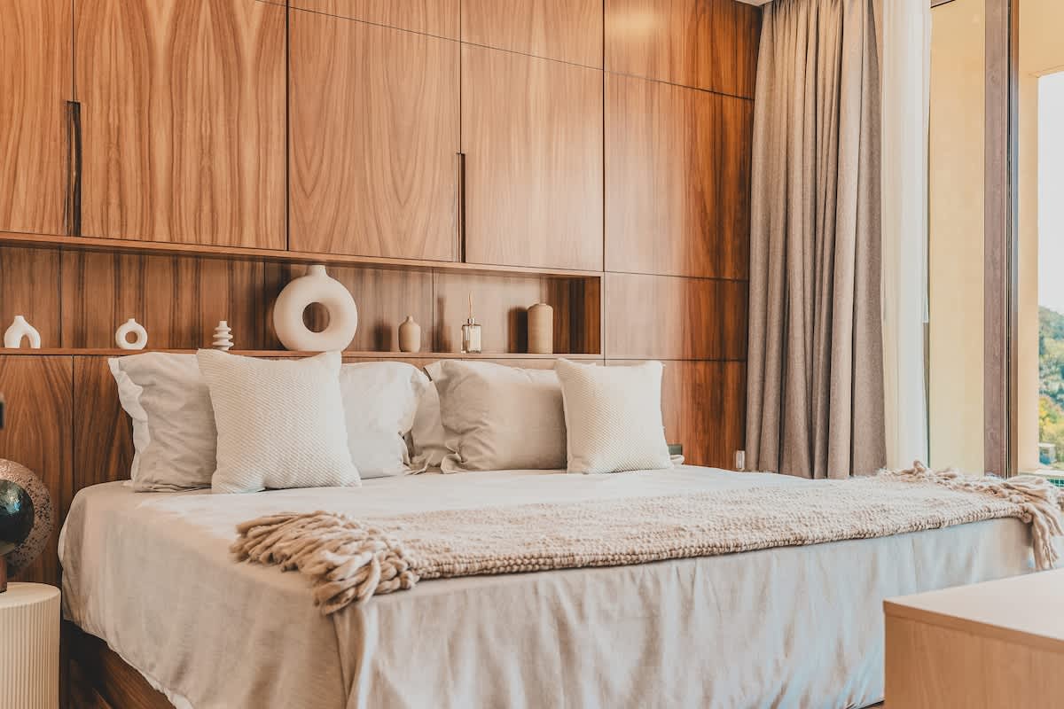 This bedroom features a large bed with white and beige bedding, flanked by a built-in wooden headboard with integrated shelving displaying decorative vases. Floor-to-ceiling curtains and a large window are visible on the right.