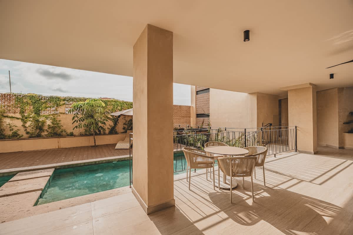 This image shows a covered outdoor patio area next to a swimming pool, furnished with a round dining table and four chairs, offering a comfortable spot for relaxation or dining by the pool. The pool deck has lounge chairs and is surrounded by greenery.