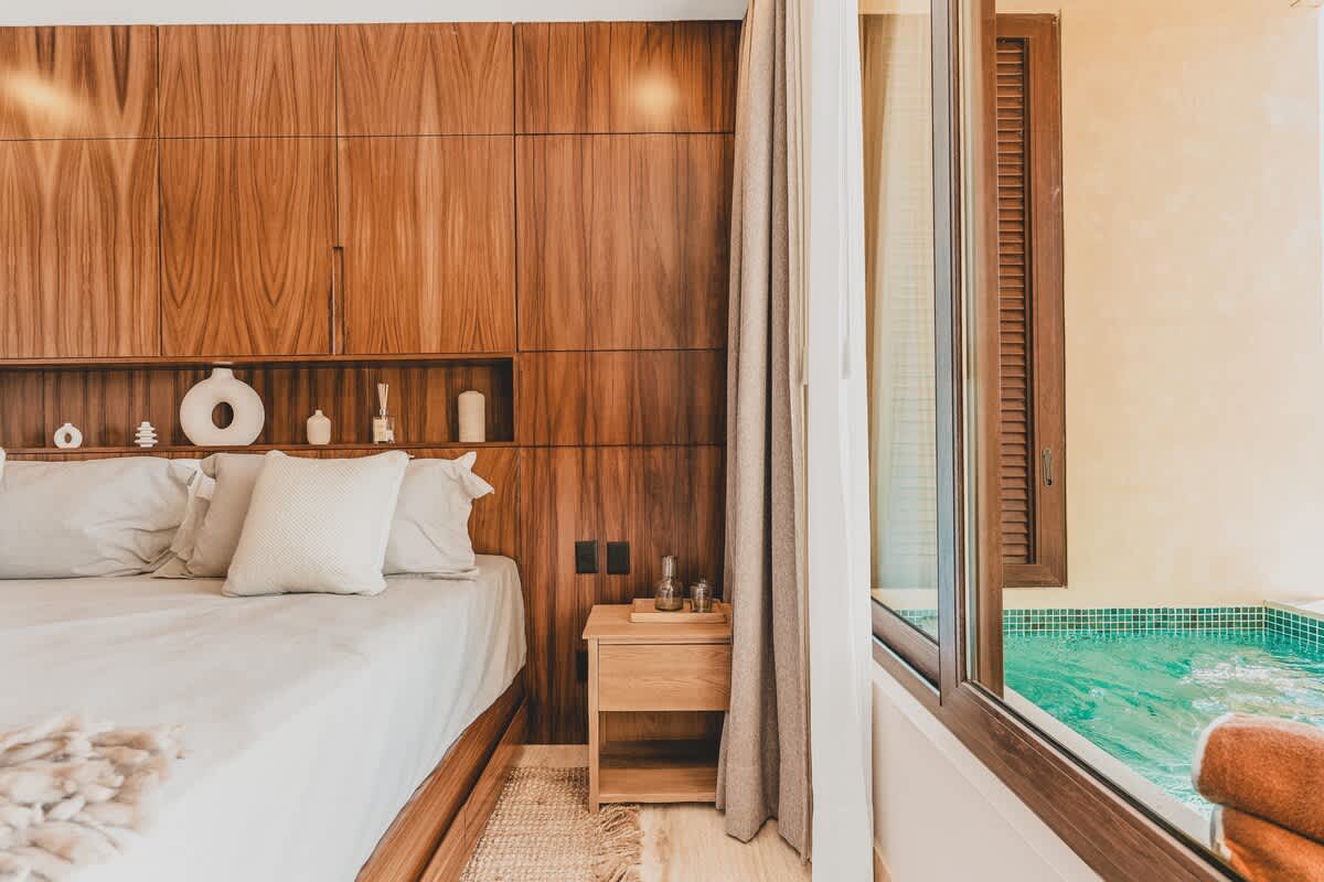 This bedroom features a large bed with neutral bedding and a wooden headboard with integrated shelving. A large window and glass door open to a private outdoor hot tub with blue mosaic tiles.