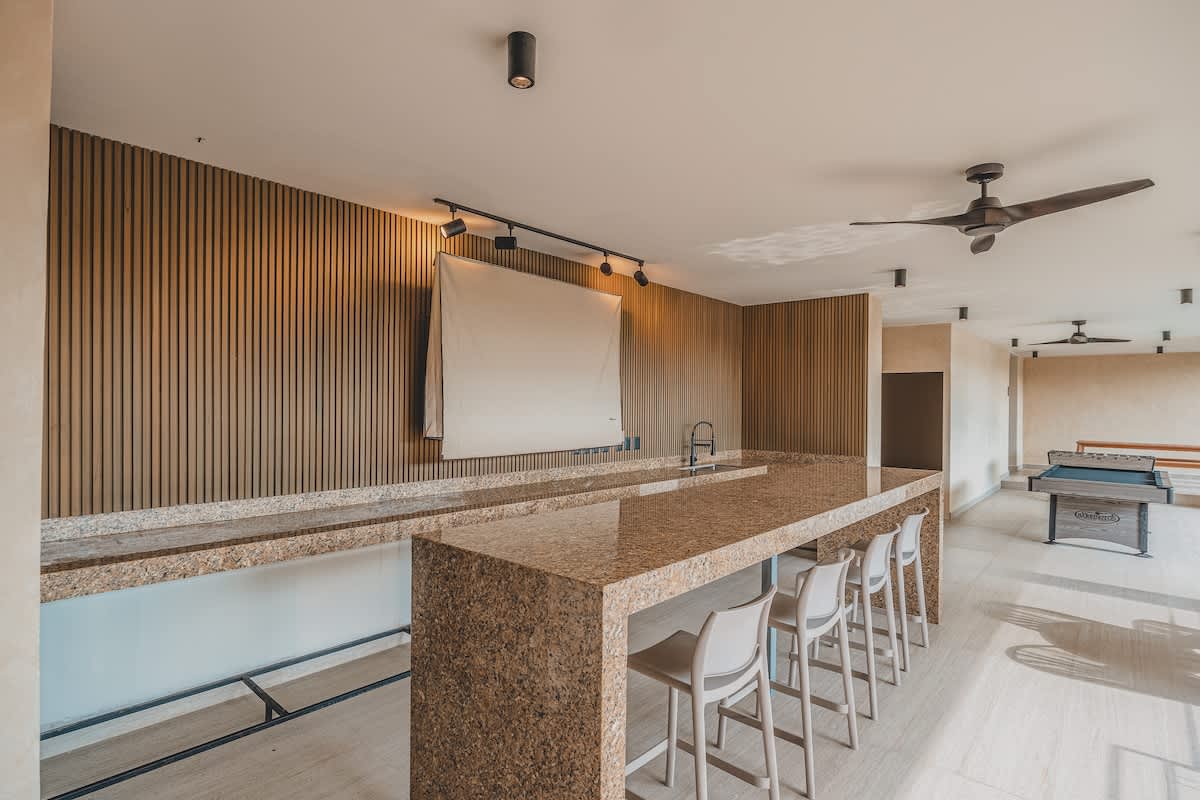 A modern common area featuring a long granite-topped bar with bar stools, wood-slat accent walls, track lighting, and a pool table visible in the background.